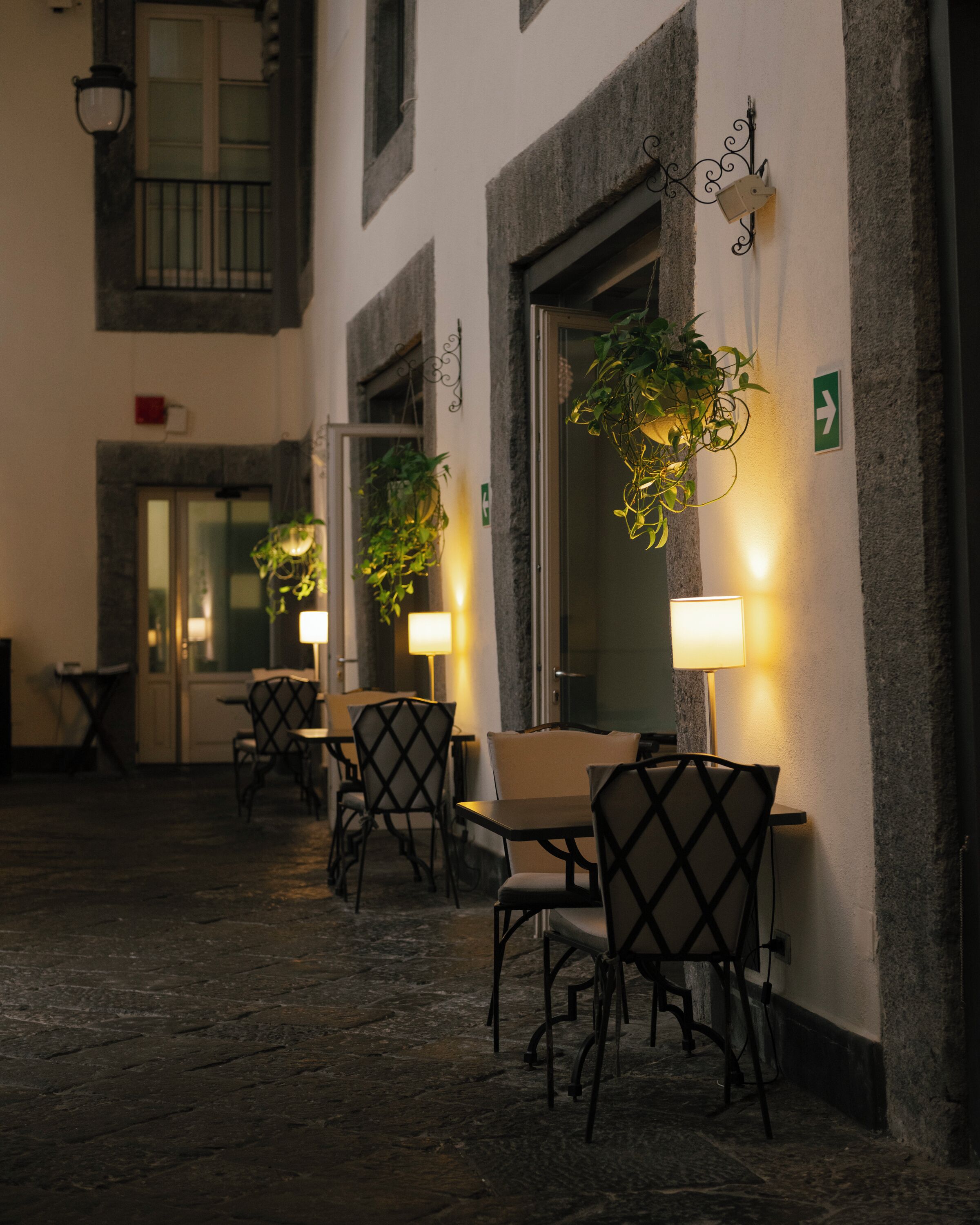 An outdoor cafe area with tables and chairs, illuminated by wall-mounted lamps, and a potted plant hanging from the wall.