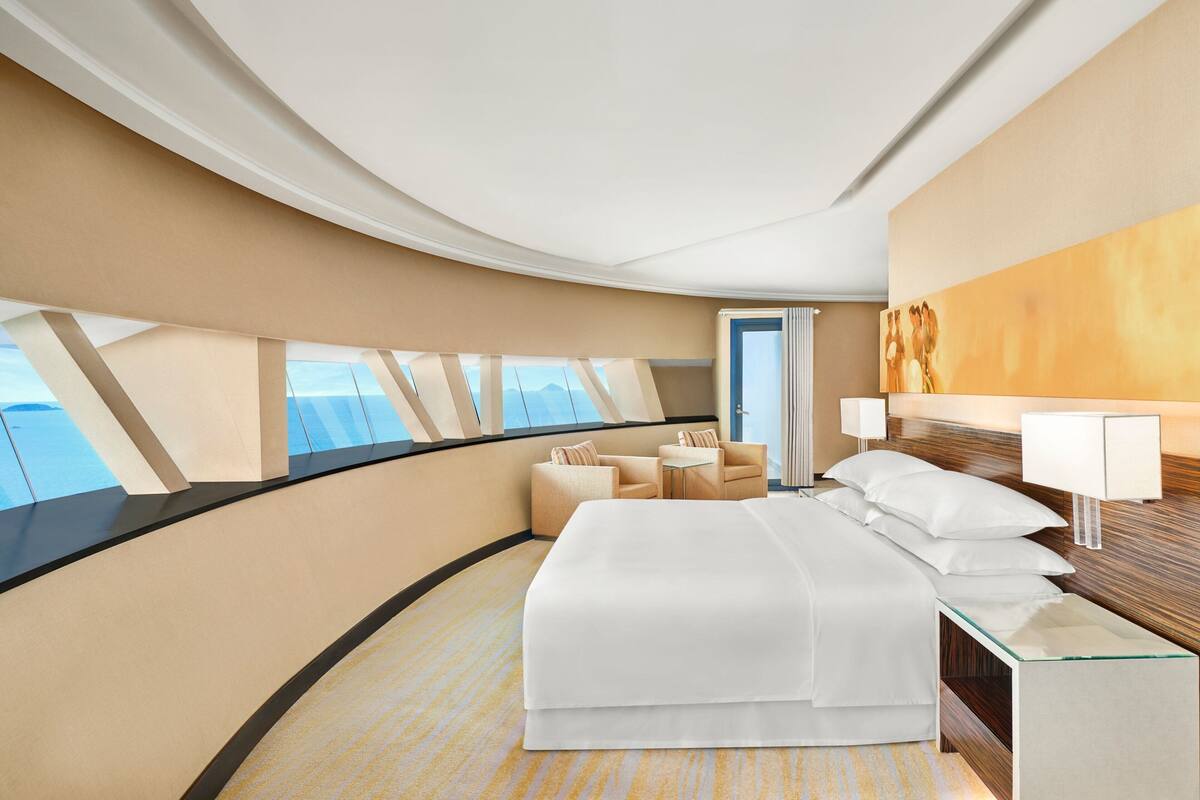 Une chambre d’hôtel moderne dotée d’un plafond incurvé, d’un grand lit, d’un coin salon avec deux fauteuils et d’une vue sur la mer par les fenêtres.
