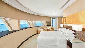 Een moderne hotelkamer met een gebogen plafond, een groot bed, een zithoek met twee stoelen en uitzicht op zee door de ramen.