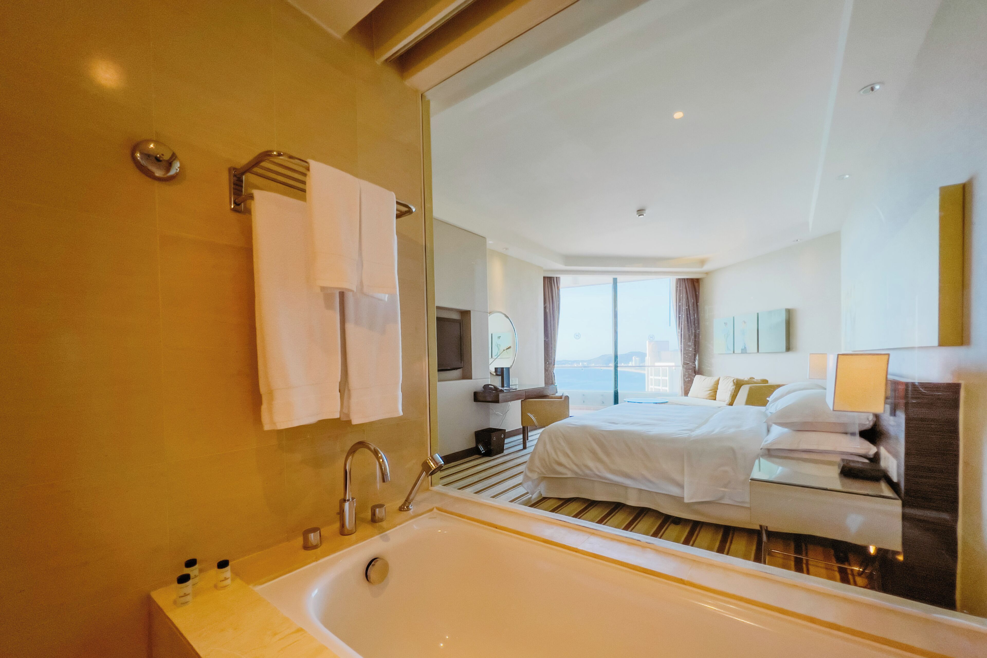 A hotel room with a large bathtub, a bed with white linens, a bedside table, and a view of the city.