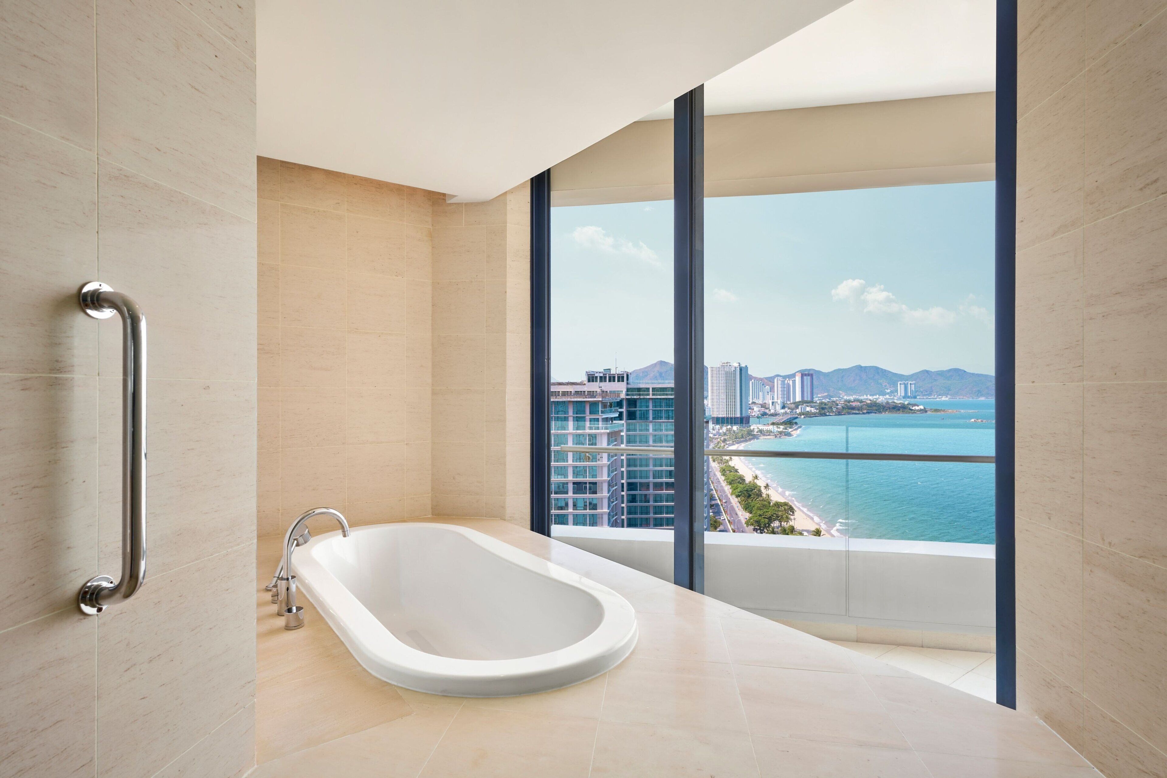A modern bathroom with a freestanding bathtub, a handrail, and a large window offering a view of a coastal cityscape.