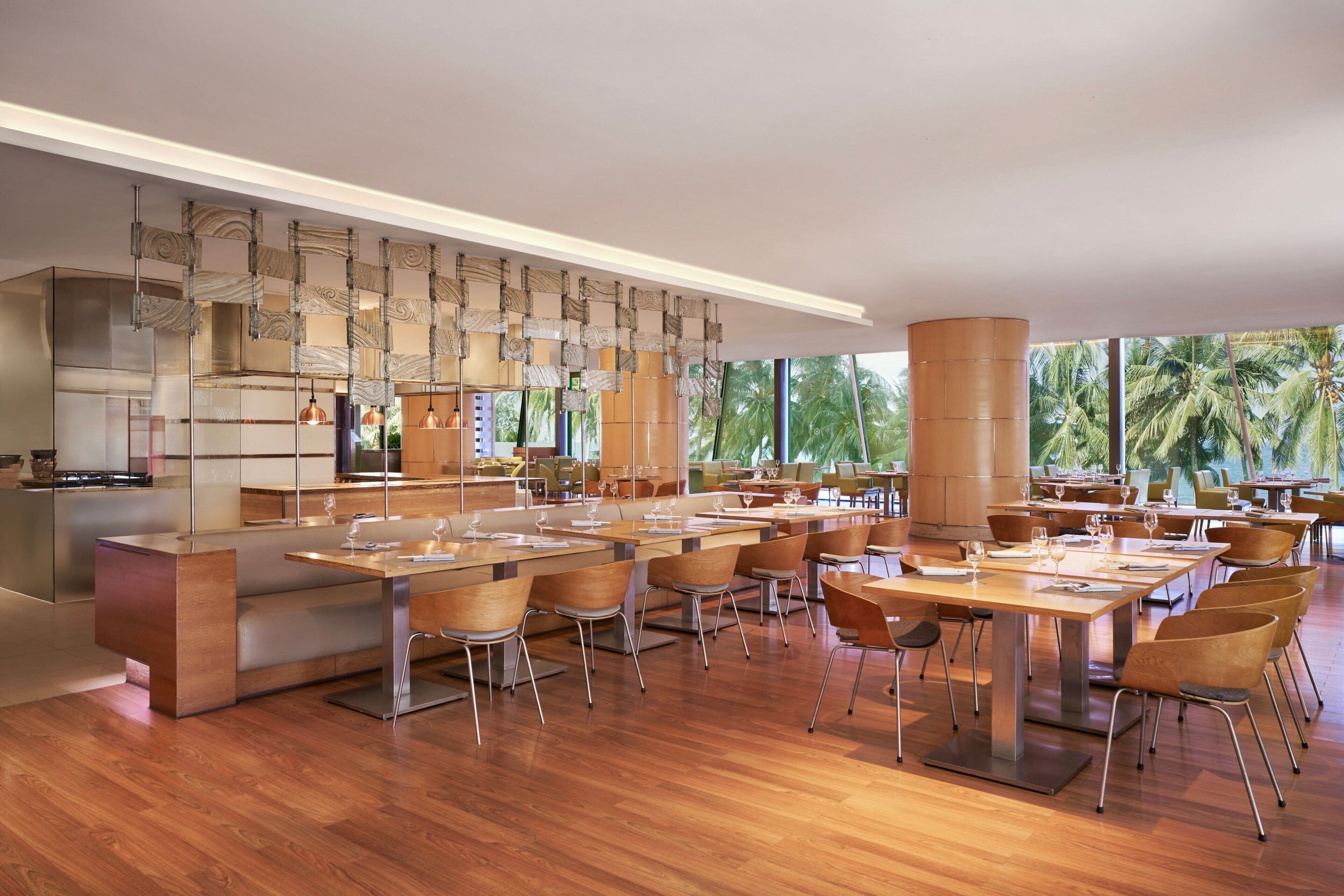 A modern dining area with wooden tables and chairs, a central bar, and large windows with a view of palm trees.