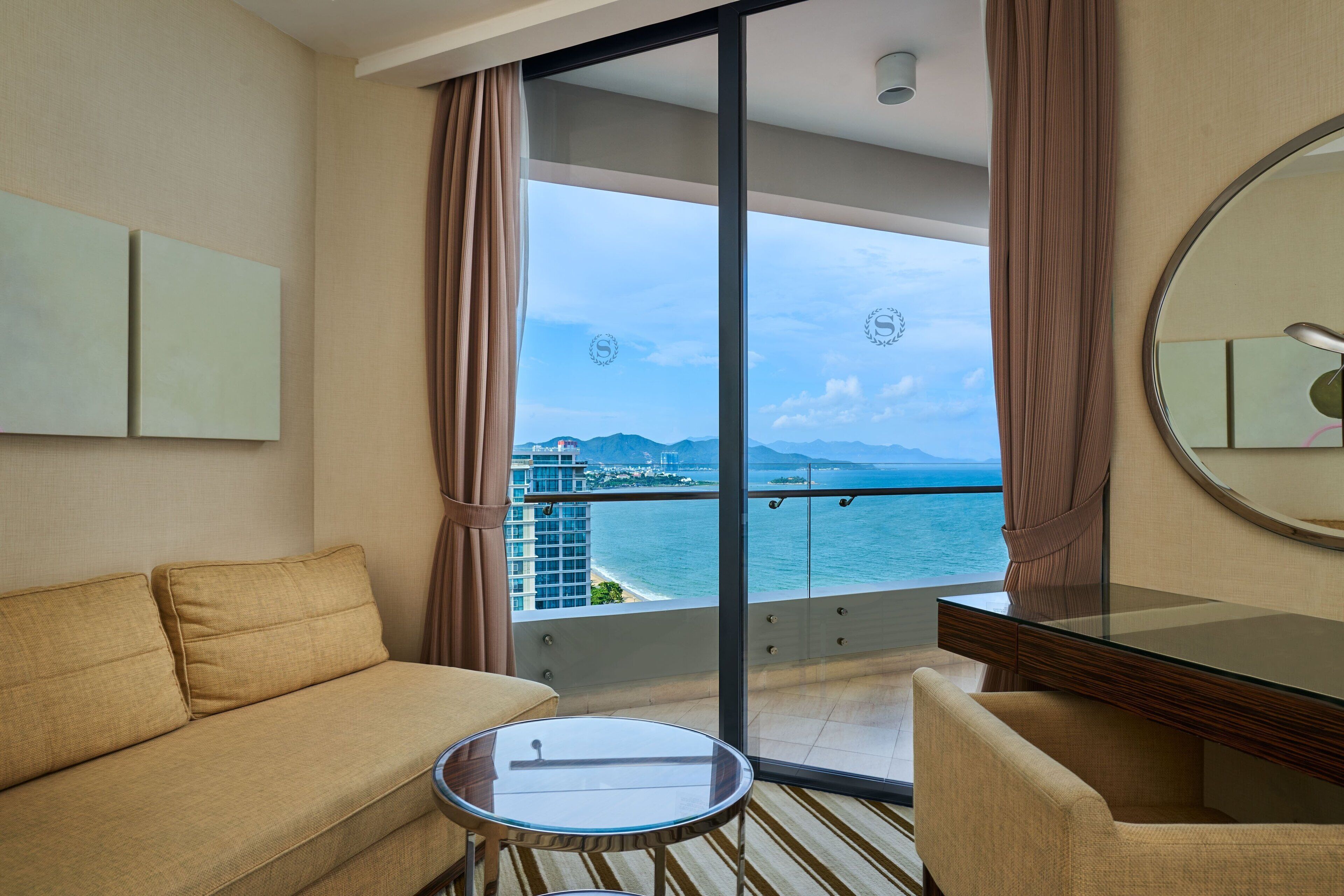 Une chambre d’hôtel avec un canapé, une petite table, un miroir et un balcon offrant une vue sur la mer et les bâtiments.