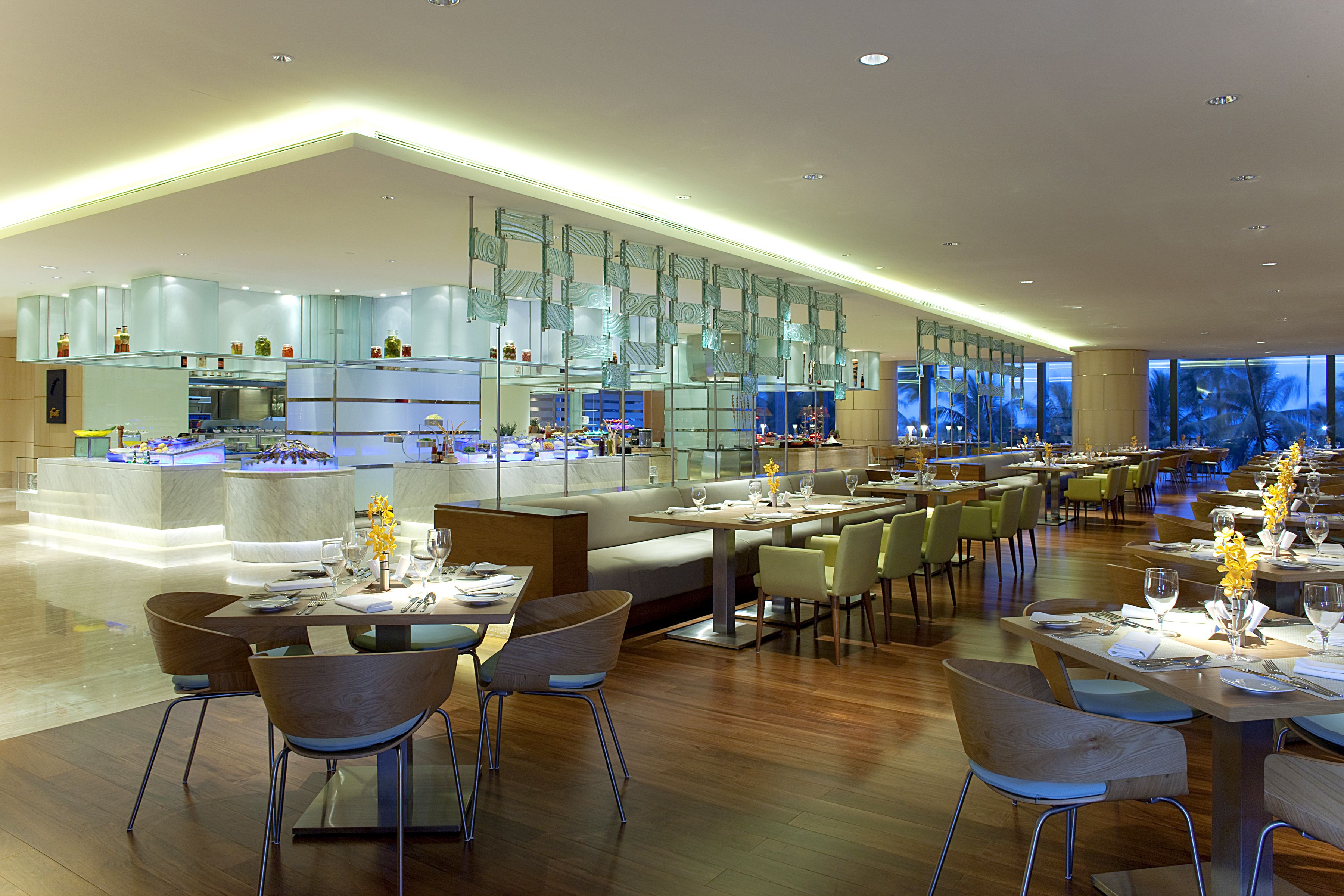 A modern restaurant with a central bar, multiple dining tables, and a large glass wall.