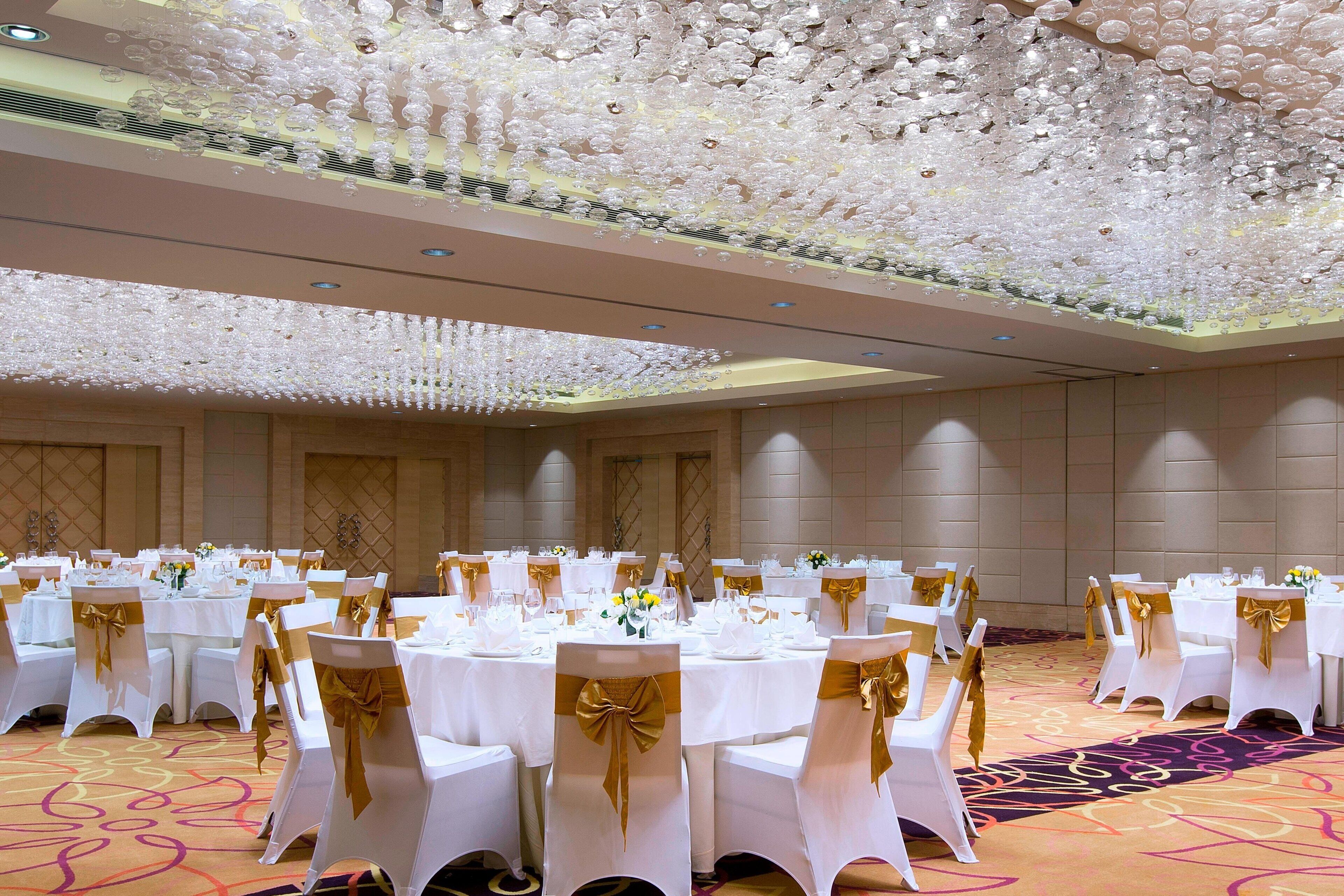A banquet hall with round tables, white chairs with gold sashes, and a ceiling adorned with numerous white spherical decorations.