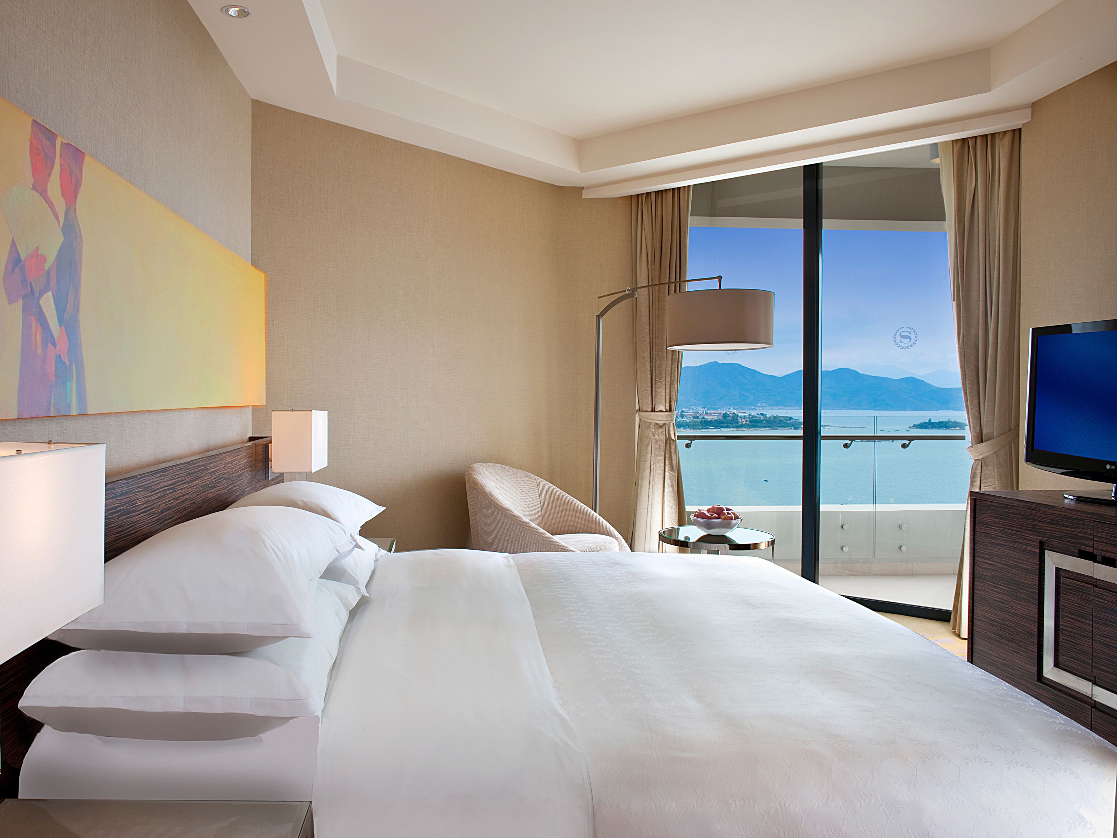 Une chambre d’hôtel avec un grand lit, une télévision, un balcon avec vue sur la mer et les montagnes, et un tableau accroché au mur.