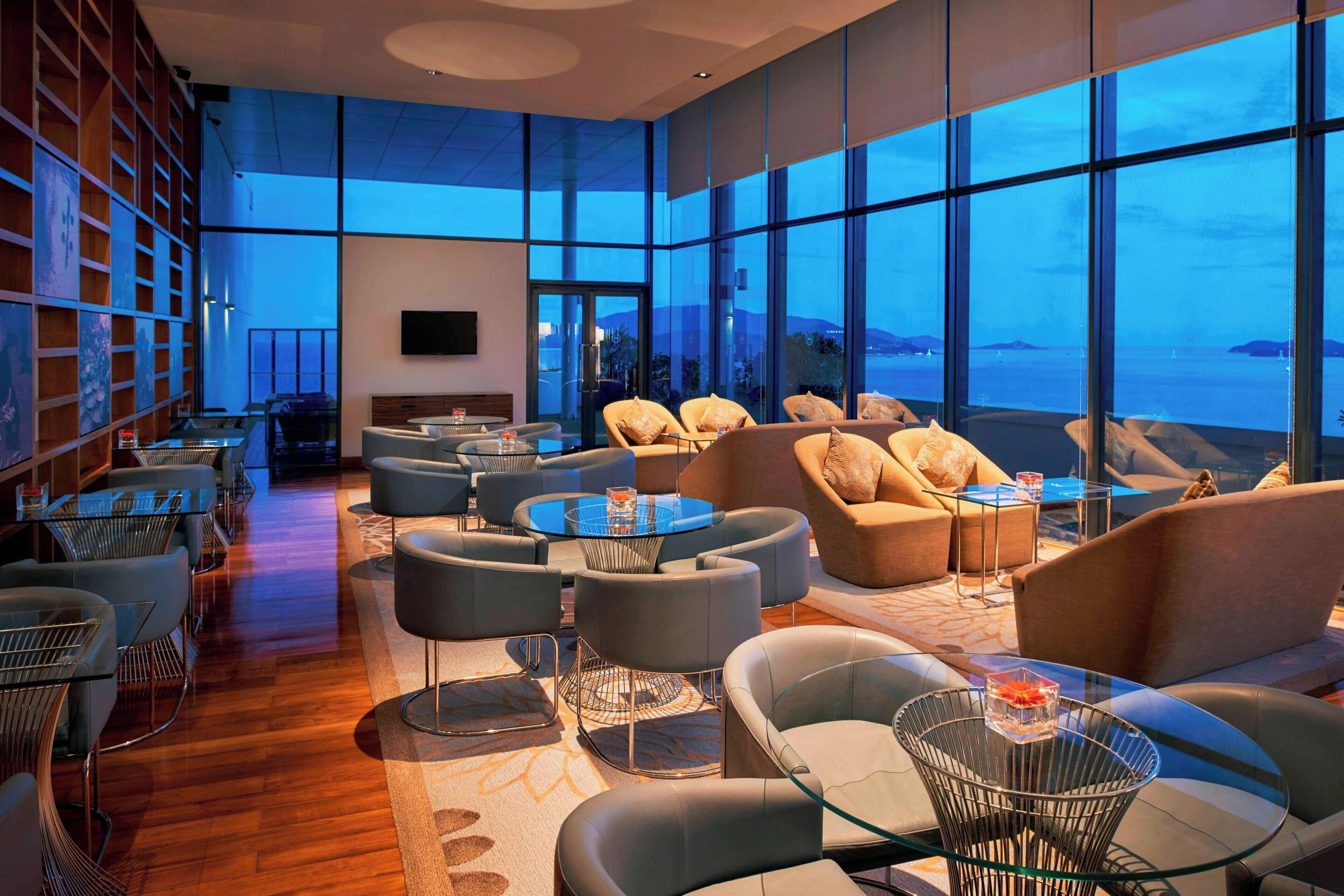 A modern lounge area with comfortable seating, a large window offering a view of the sea, and a bookshelf with various items.