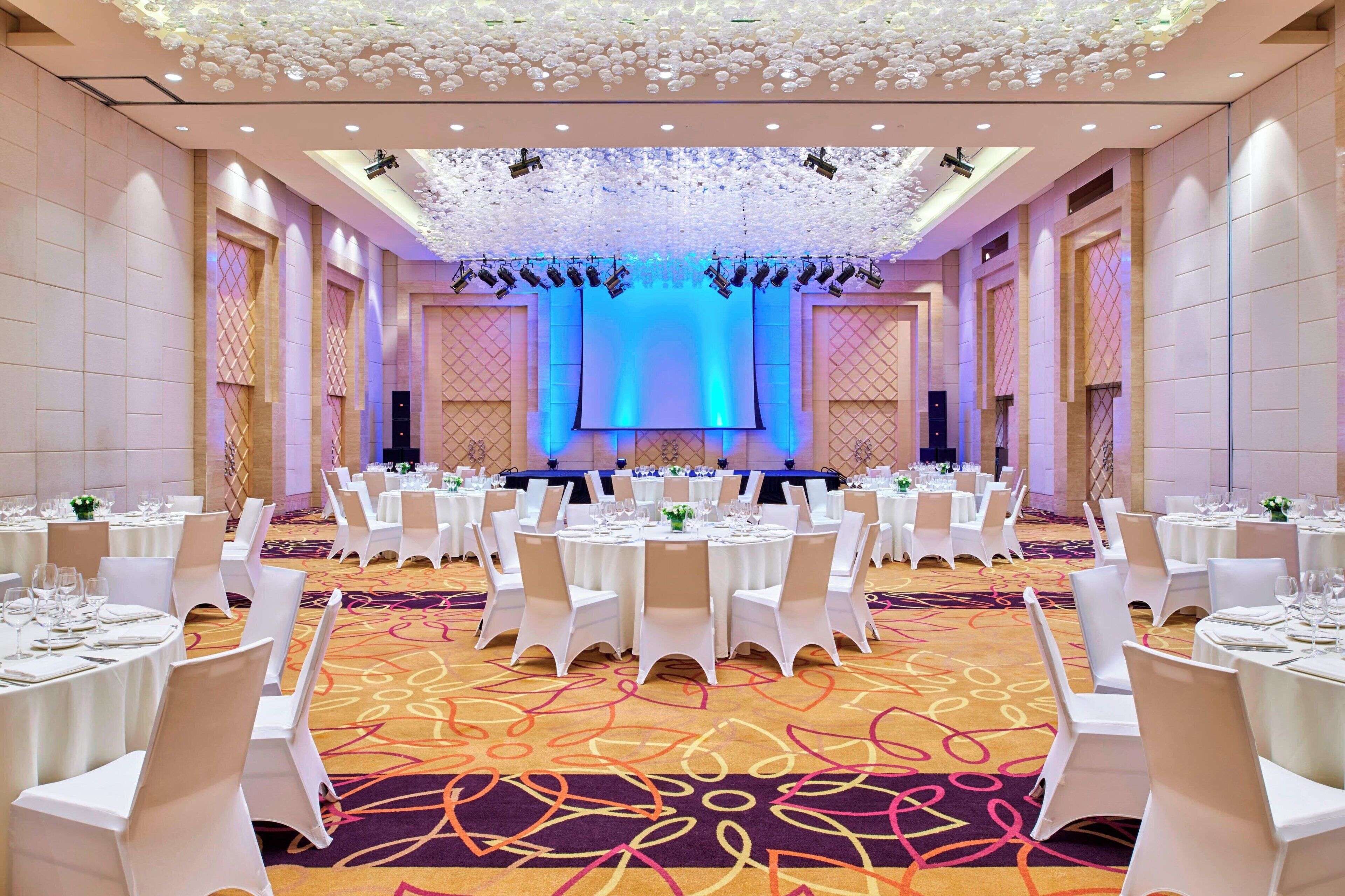 A banquet hall with round tables, white chairs, and a stage with blue lighting.