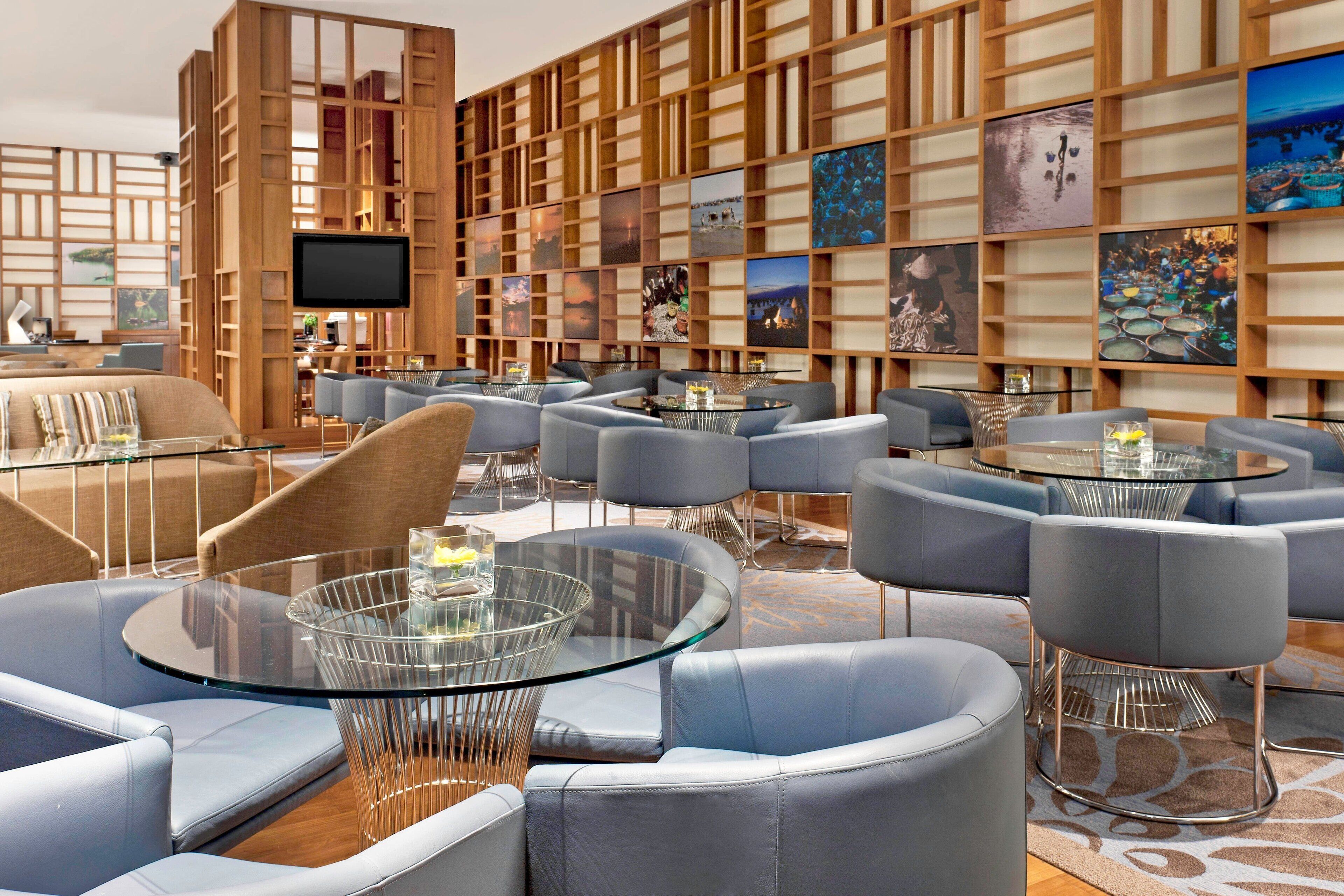 A modern lounge area with round glass tables, grey upholstered sofas, and a wooden wall with artwork.