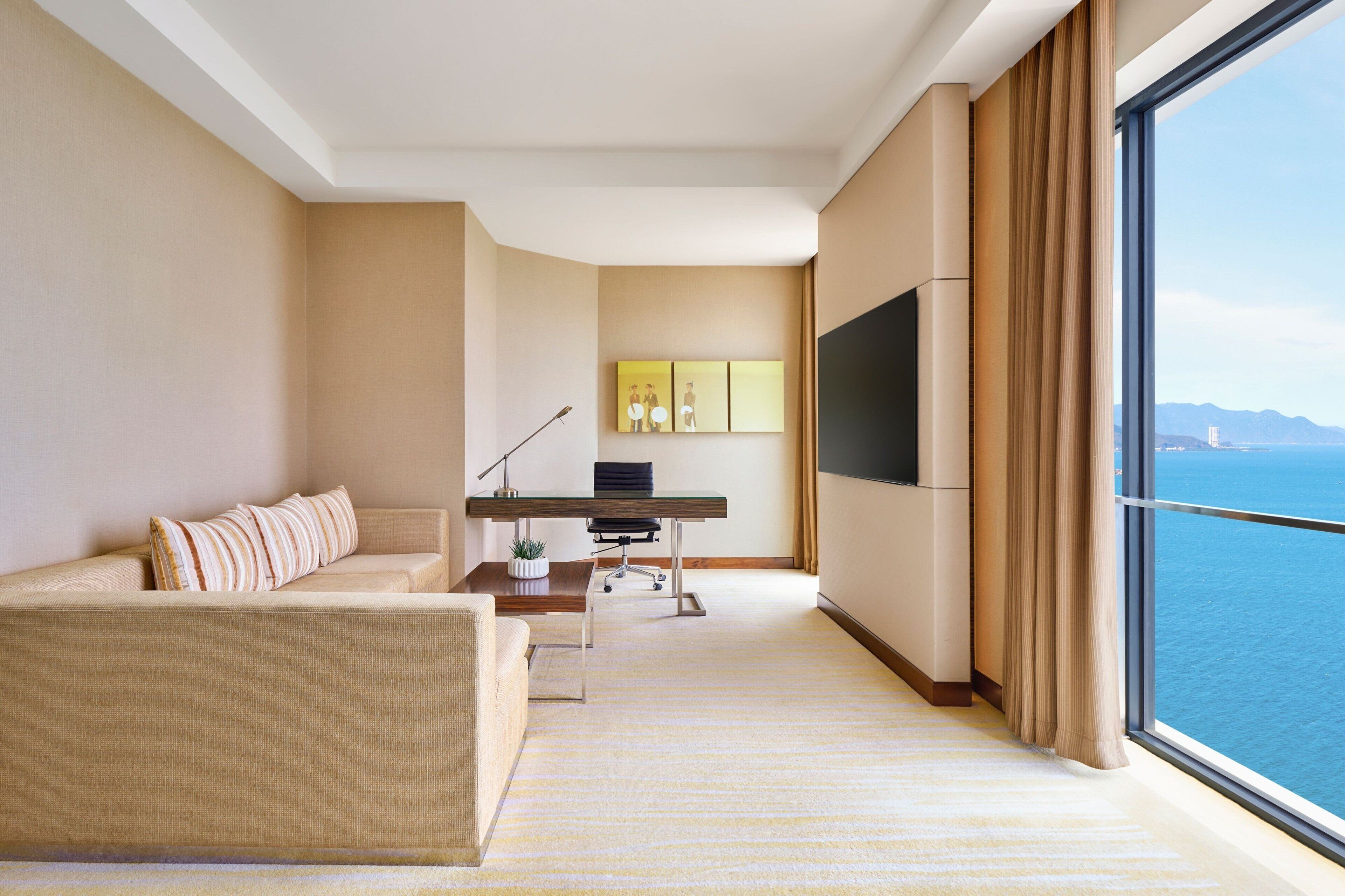 Habitación de hotel moderna con un ventanal grande, un sofá, un escritorio con computadora y un televisor de pantalla plana.
