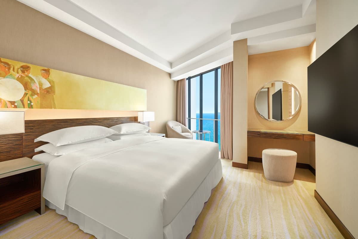 Une chambre d’hôtel moderne dotée d’un grand lit, d’un bureau avec une chaise, d’un miroir rond et offrant une vue sur l’océan grâce à de grandes fenêtres.