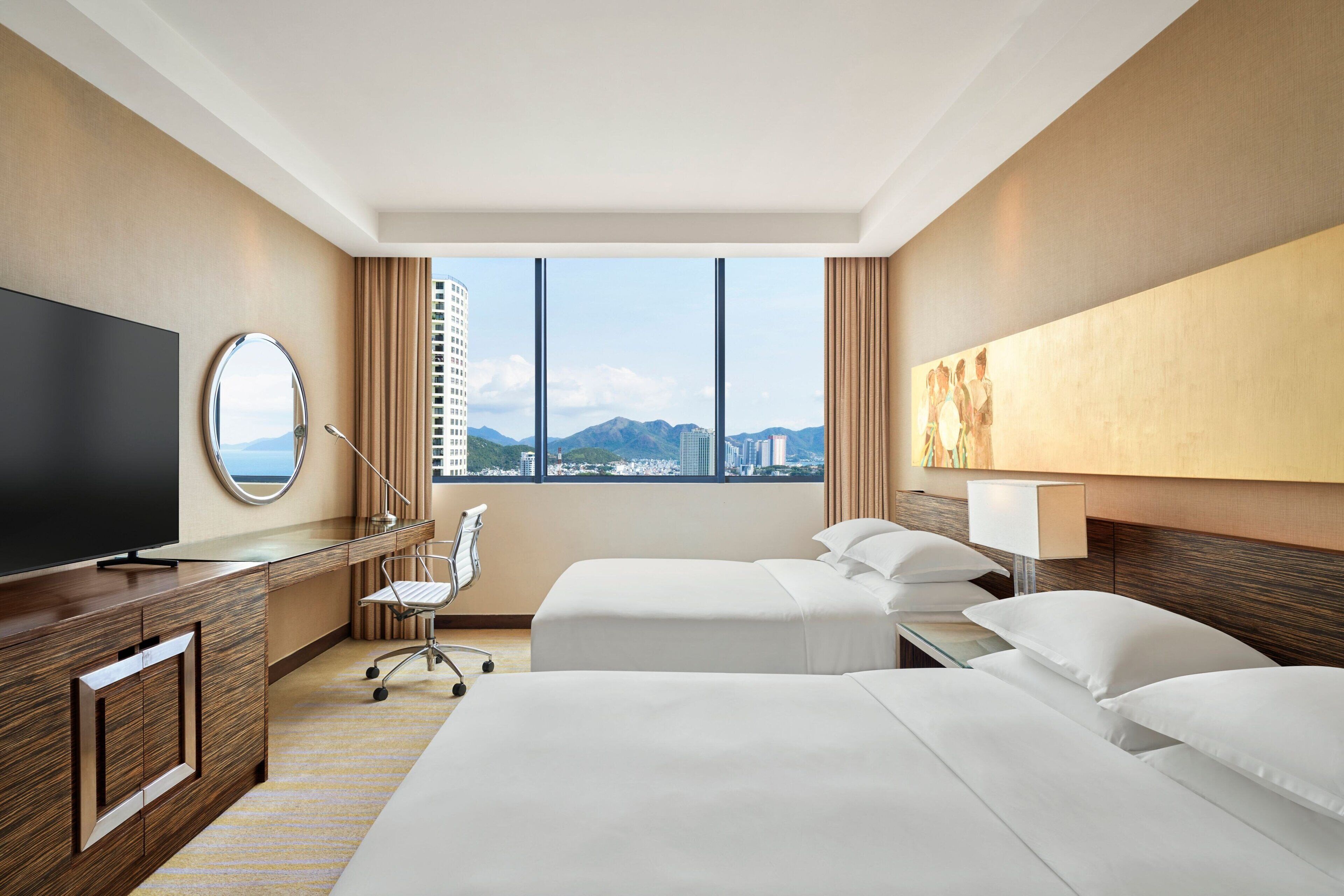 A hotel room with a large bed, a desk with a TV, a chair, and a view of the cityscape.