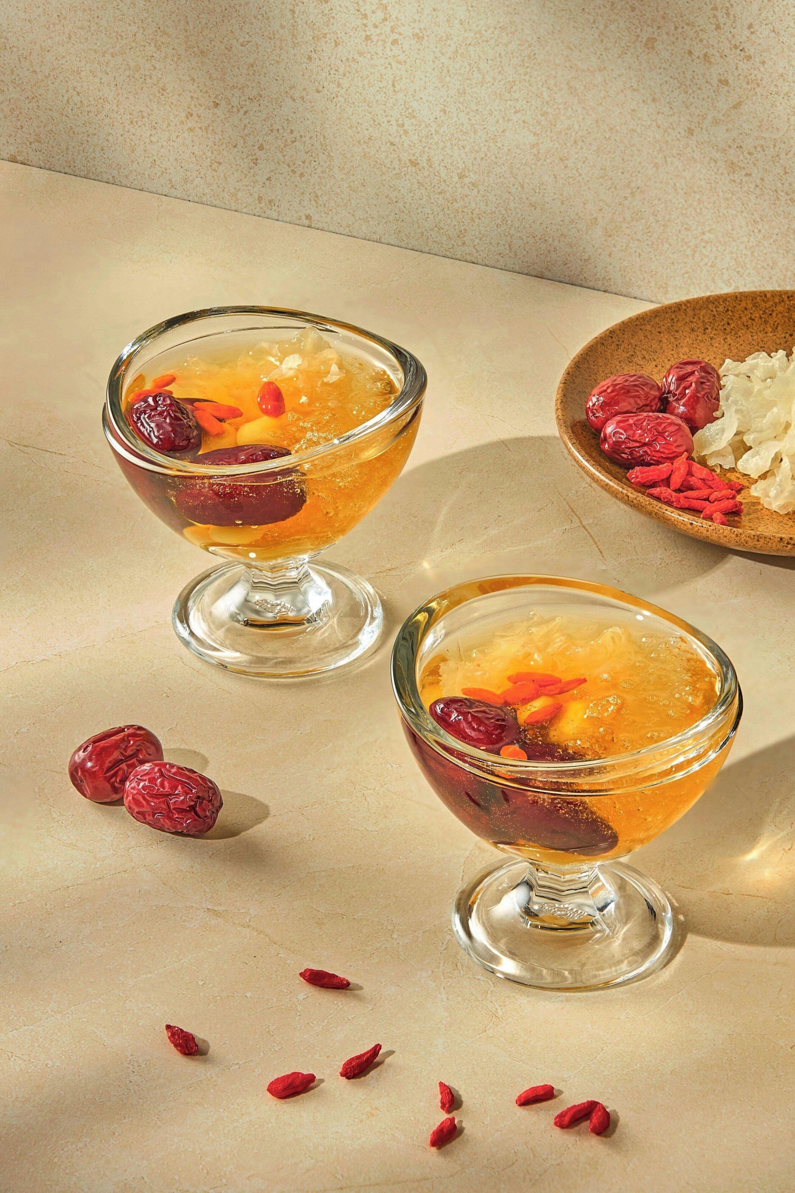 Two clear glass cups with a golden liquid and red berries on a textured surface.