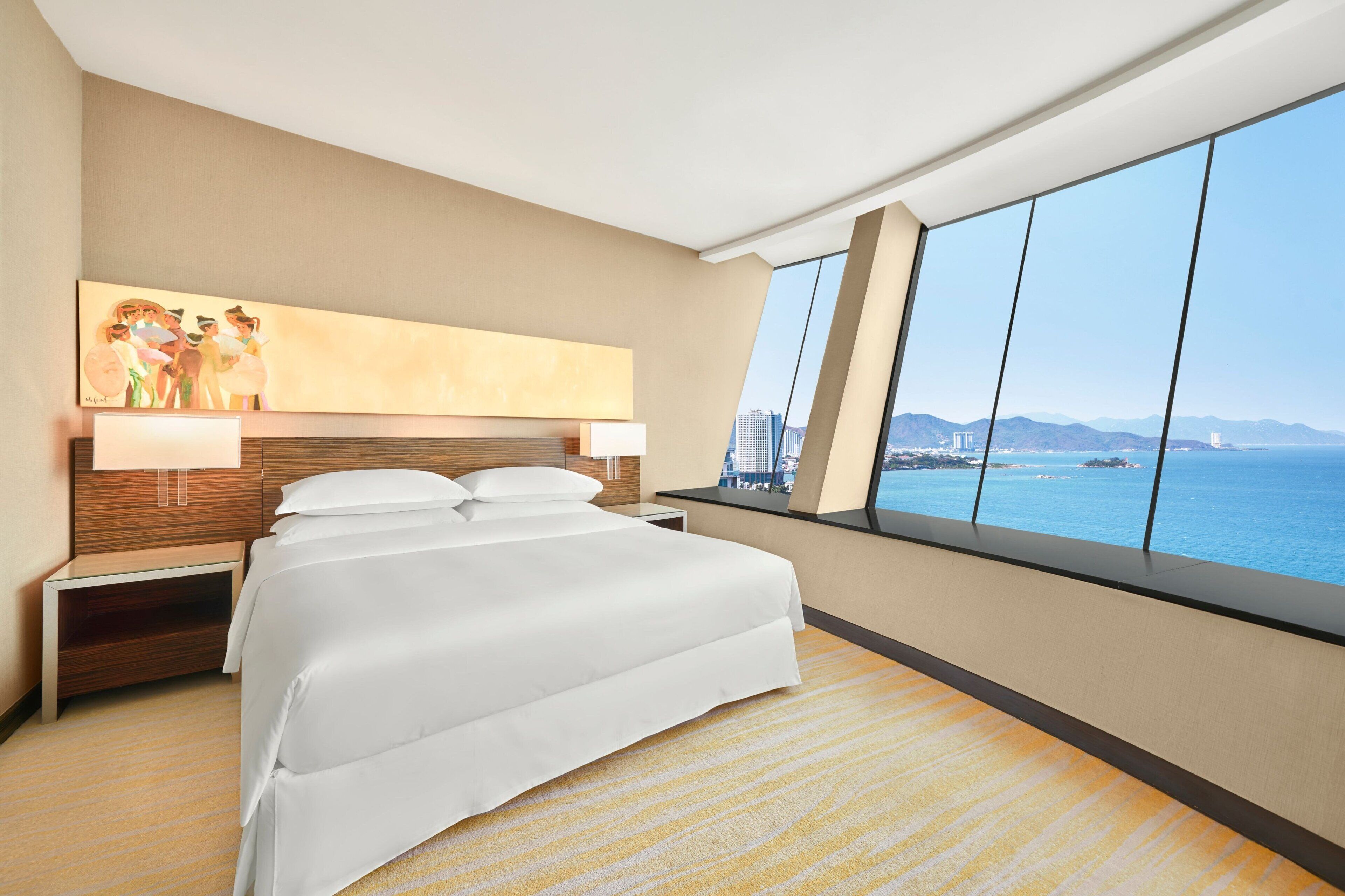 A hotel room with a large bed, a nightstand, a painting on the wall, and a view of the sea and cityscape through large windows.