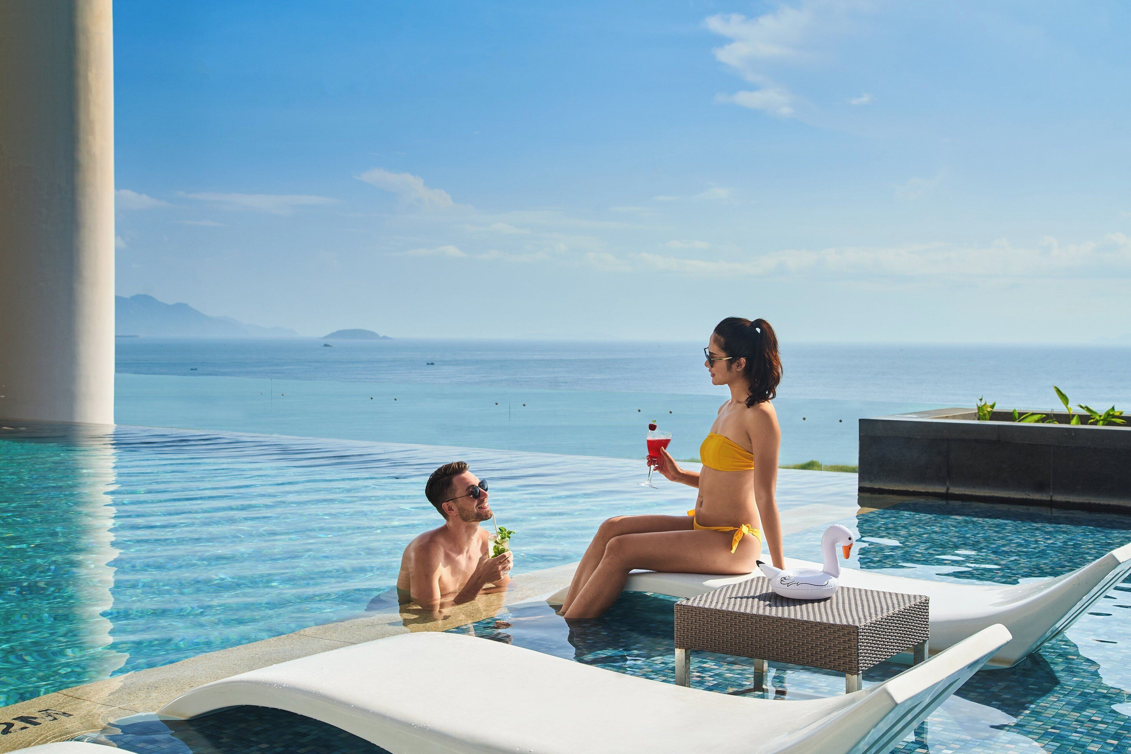 Una pareja en una piscina con un flotador de cisne, una bebida y vistas al mar.