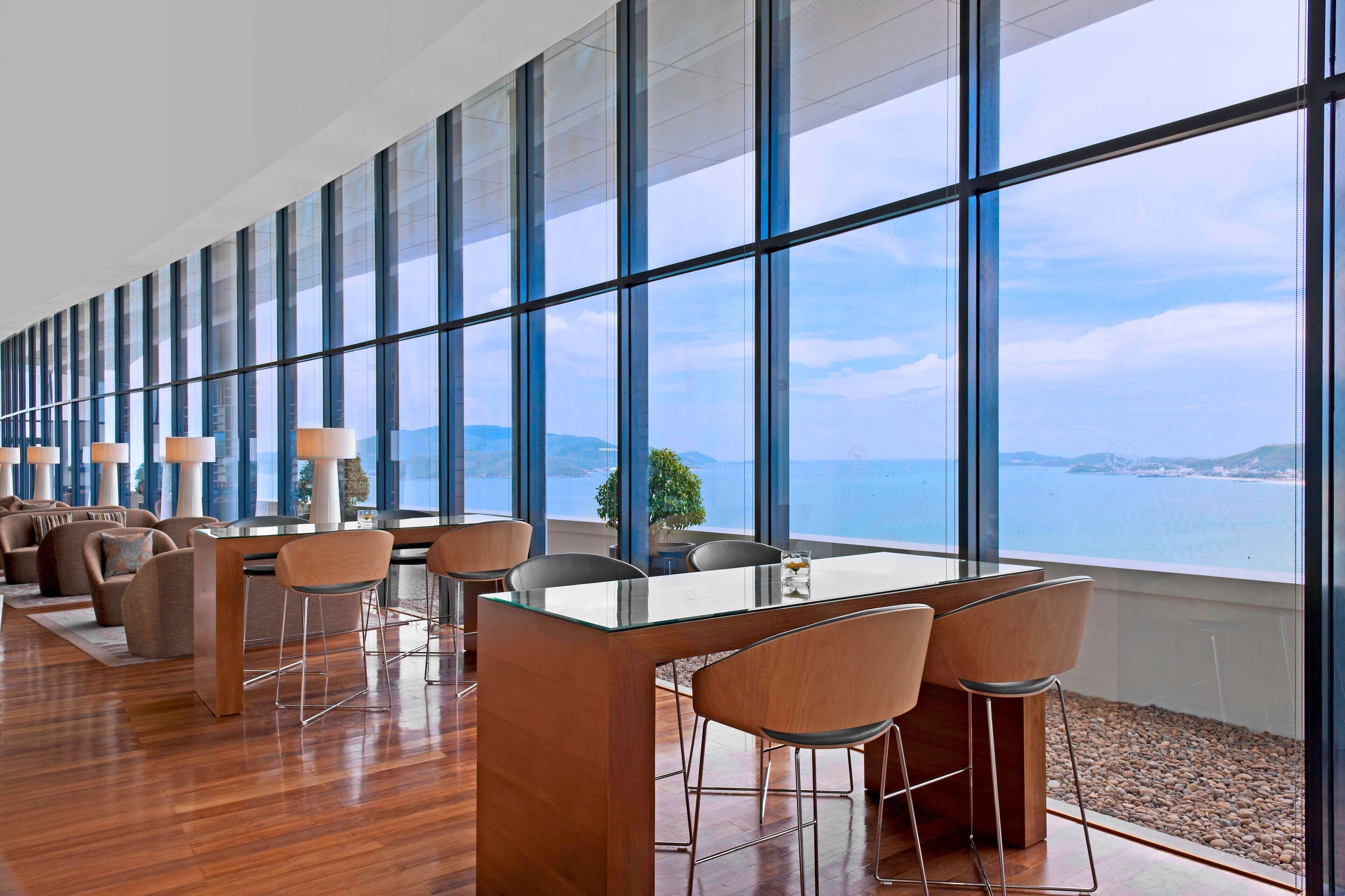 A modern dining area with a glass table and chairs, large windows offering a view of the sea and distant land.