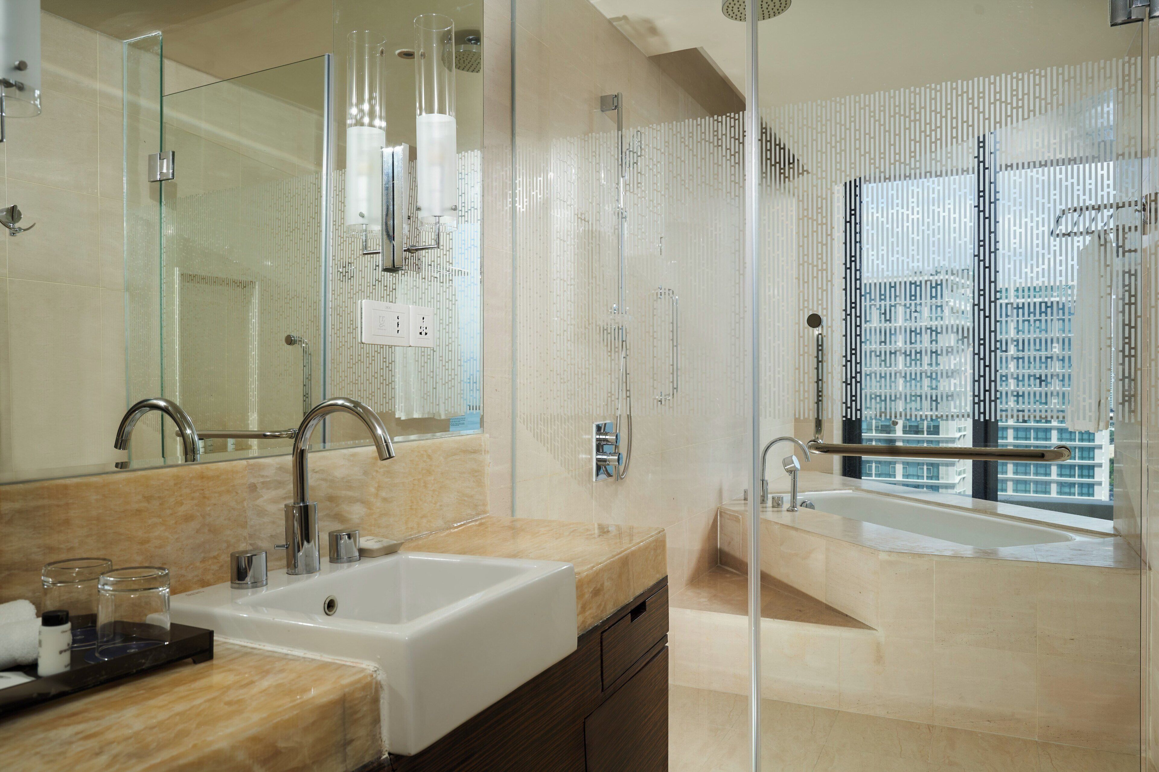 A modern bathroom with a large mirror, two sinks, a glass shower enclosure, and a view of tall buildings.