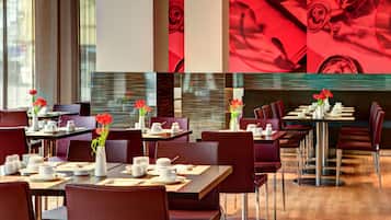 Un espace repas moderne, avec des tables préparées pour le petit-déjeuner, agrémenté de fleurs rouges dans des vases et d’une grande œuvre d’art abstraite rouge accrochée au mur.