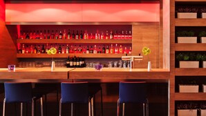 Una zona de bar con estantes de madera llenos de botellas, una barra y sillas azules.