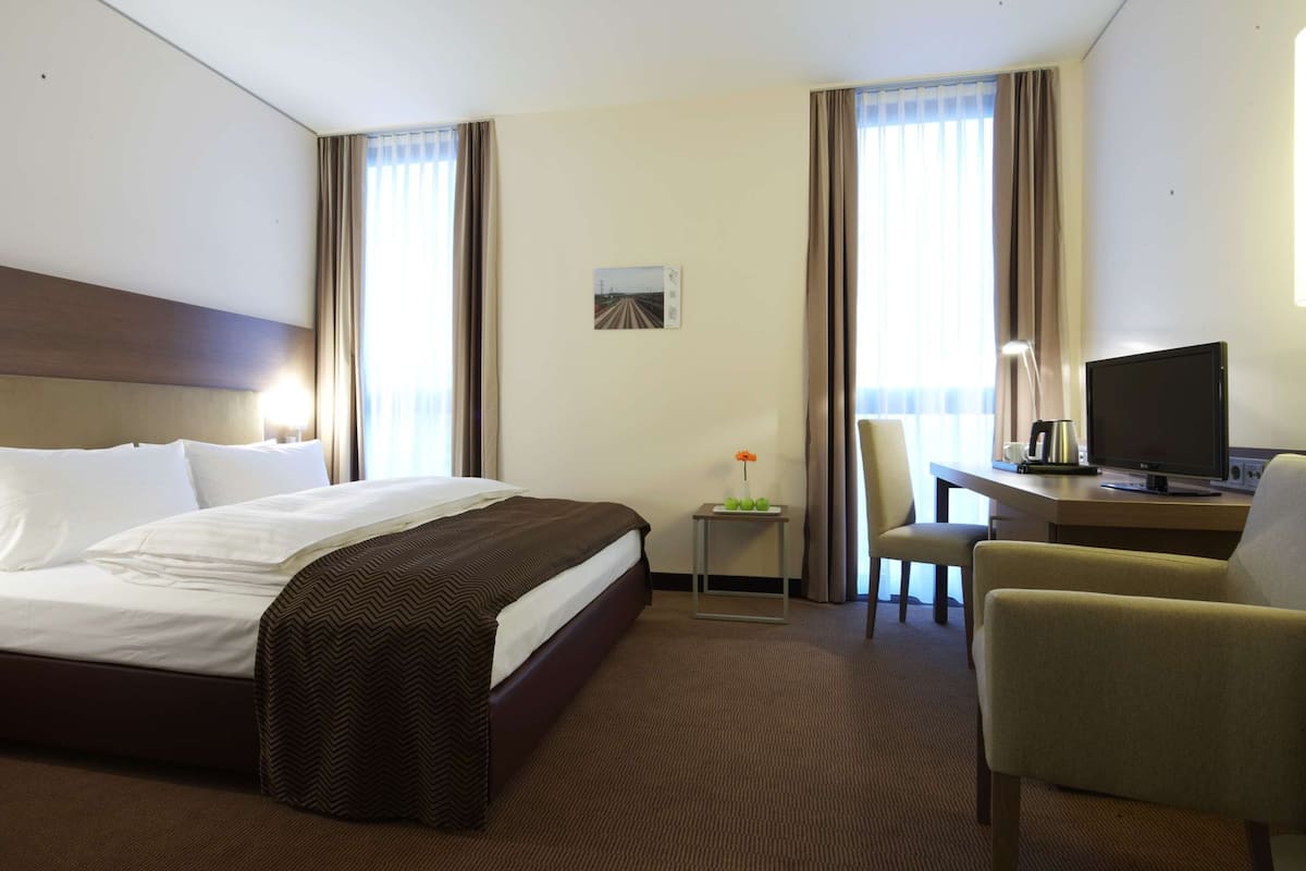 Ein Hotelzimmer mit einem großen Bett, einem Schreibtisch mit Computer, einem Sessel und einem Fernseher.