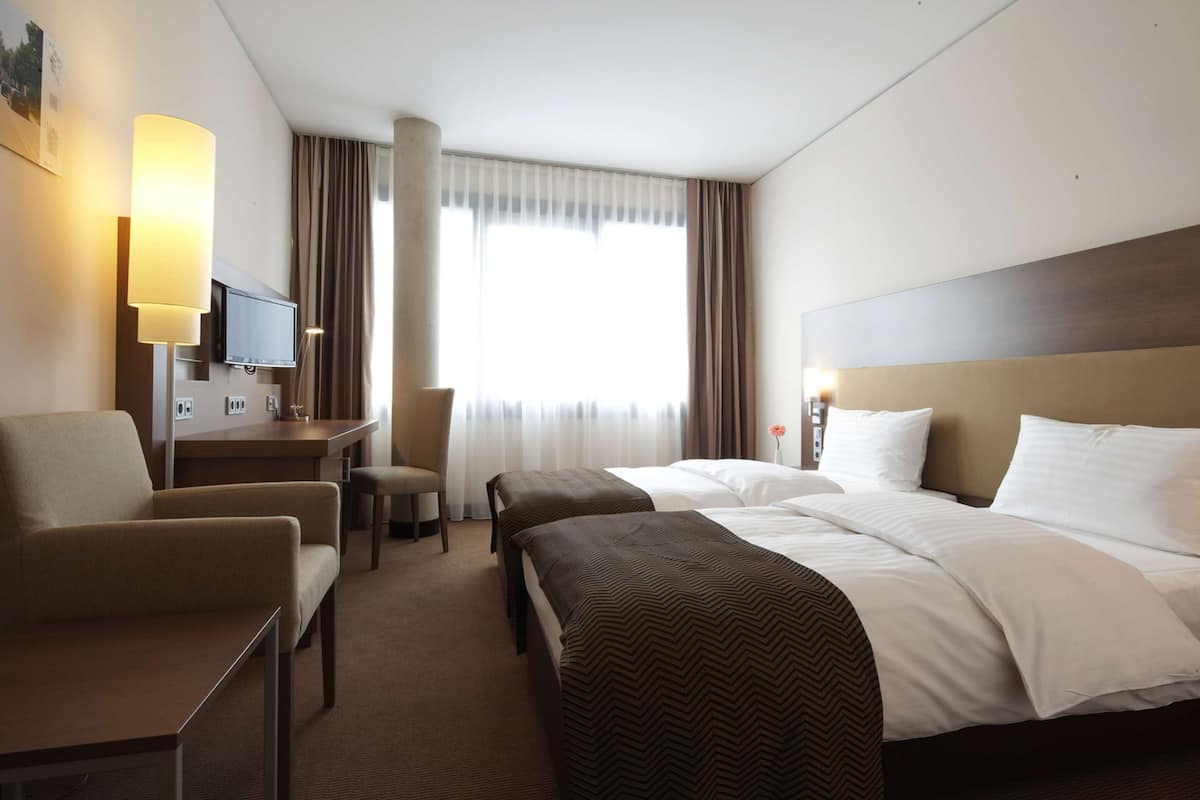 Ein Hotelzimmer mit zwei Betten, einem Schreibtisch, einem Stuhl, einem Fernseher und einem großen Fenster mit Vorhängen.