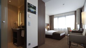 Habitación de hotel con cama, escritorio, silla, baño con ducha y una ventana grande con cortinas.