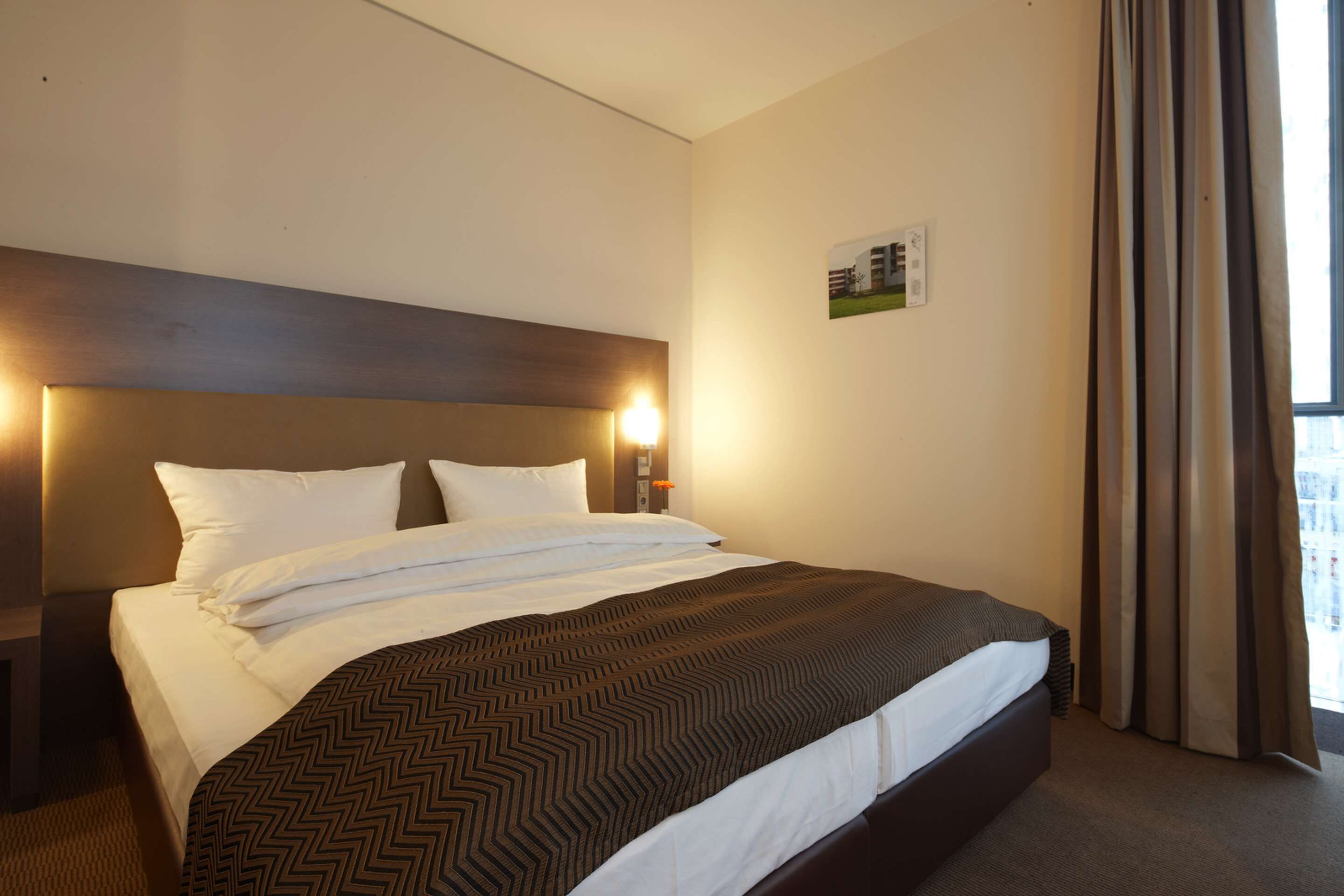 A hotel room with a large bed, a headboard with integrated lighting, a nightstand, and a window with curtains.