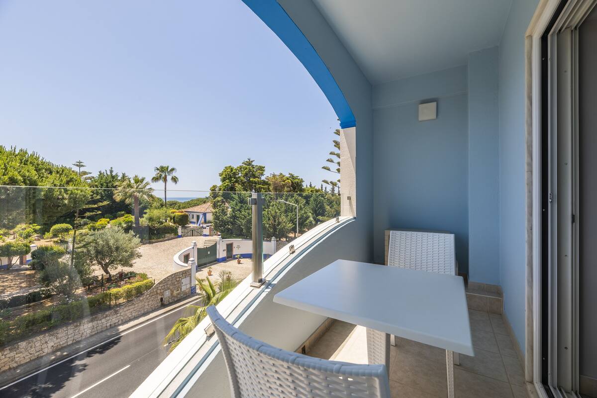 A balcony with a white table and chair, overlooking a road and greenery.
