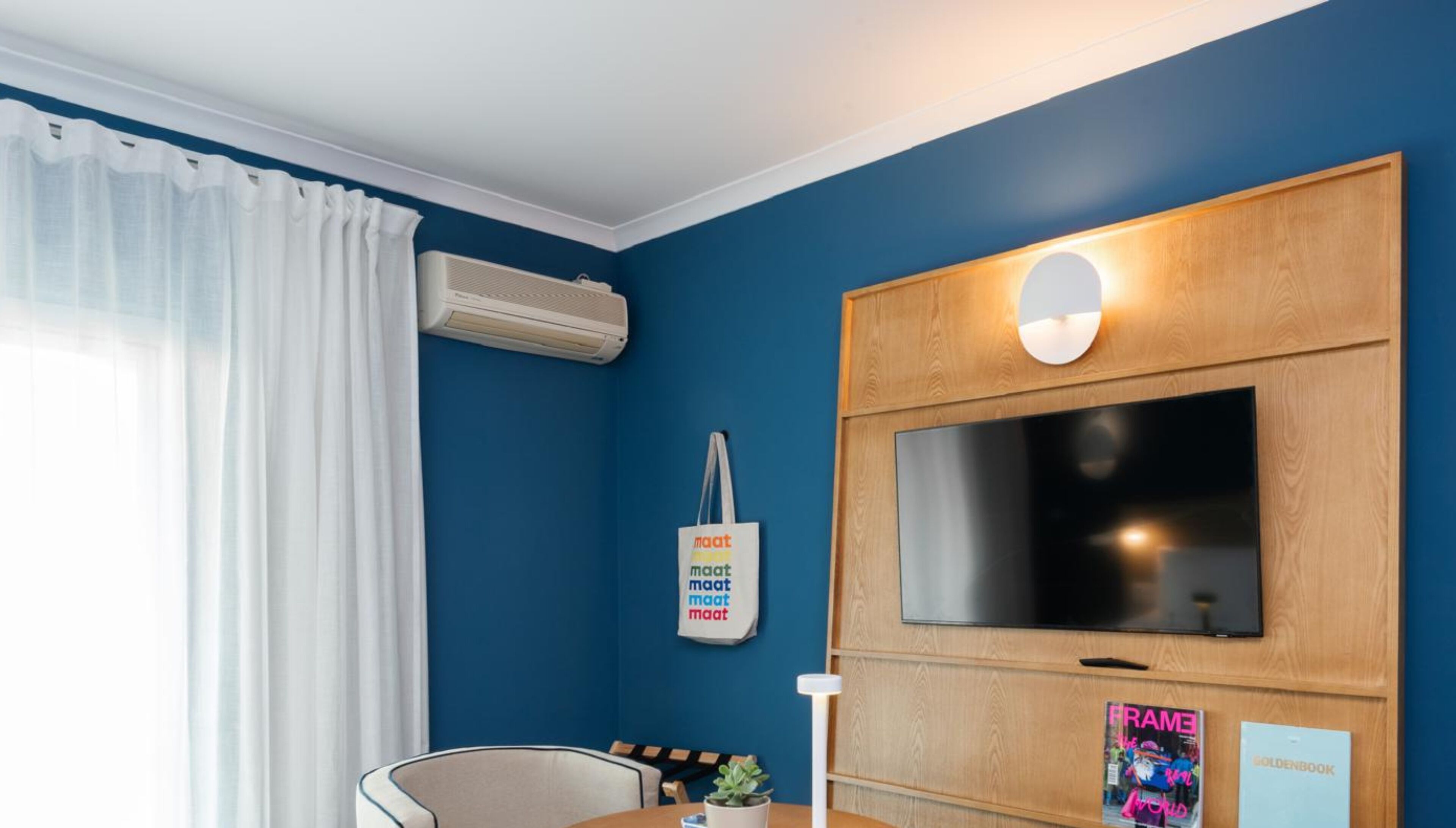 Una habitación con pared azul, televisor montado, unidad de aire acondicionado blanca, cortina blanca, puerta de madera y lámpara fijada a la pared.