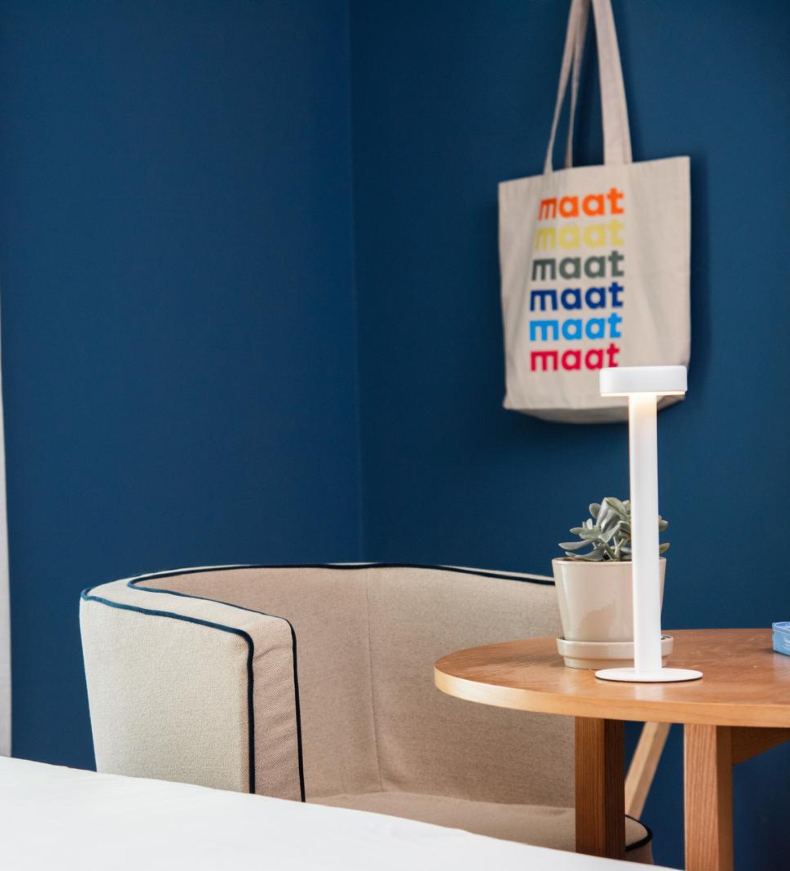 A room with a blue wall, a beige chair, a small table with a white lamp, and a potted plant.