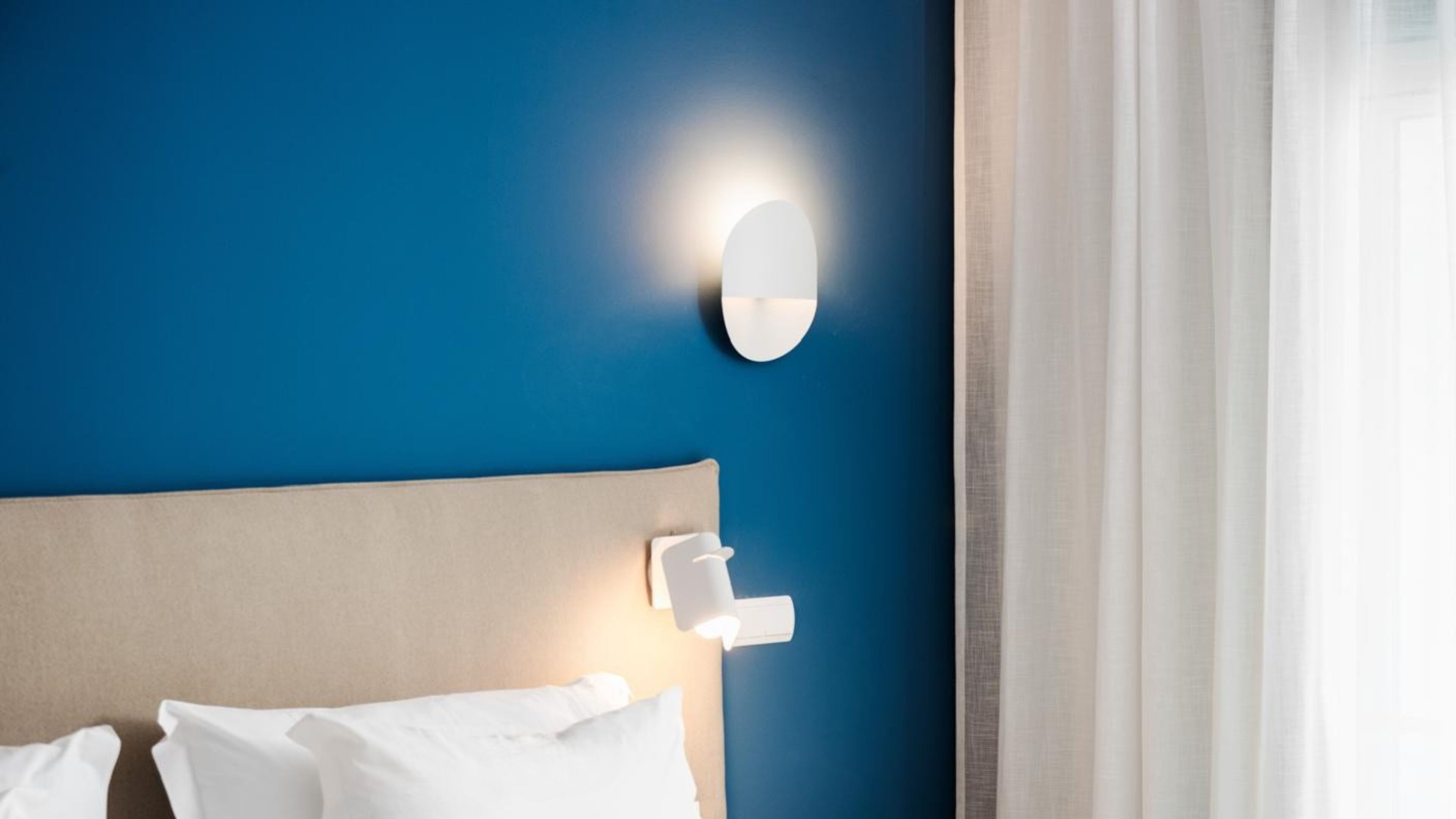 A hotel room with a blue wall, a white headboard, and a bedside lamp.