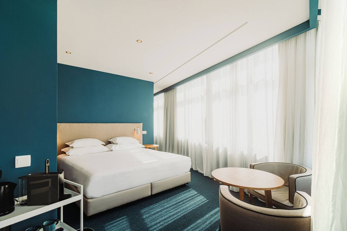 A modern hotel room with a large bed, a desk with a computer, a chair, and a small table.