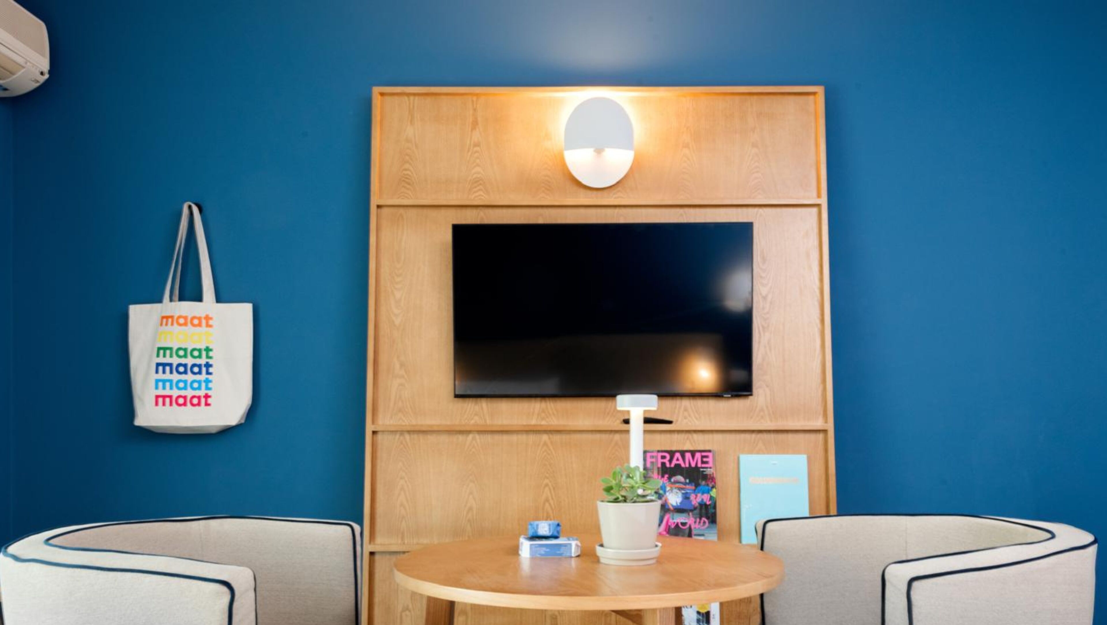 A room with a large flat-screen TV mounted on a wooden panel, a round wooden table with a plant and a magazine, two armchairs, and a wall-mounted light fixture.