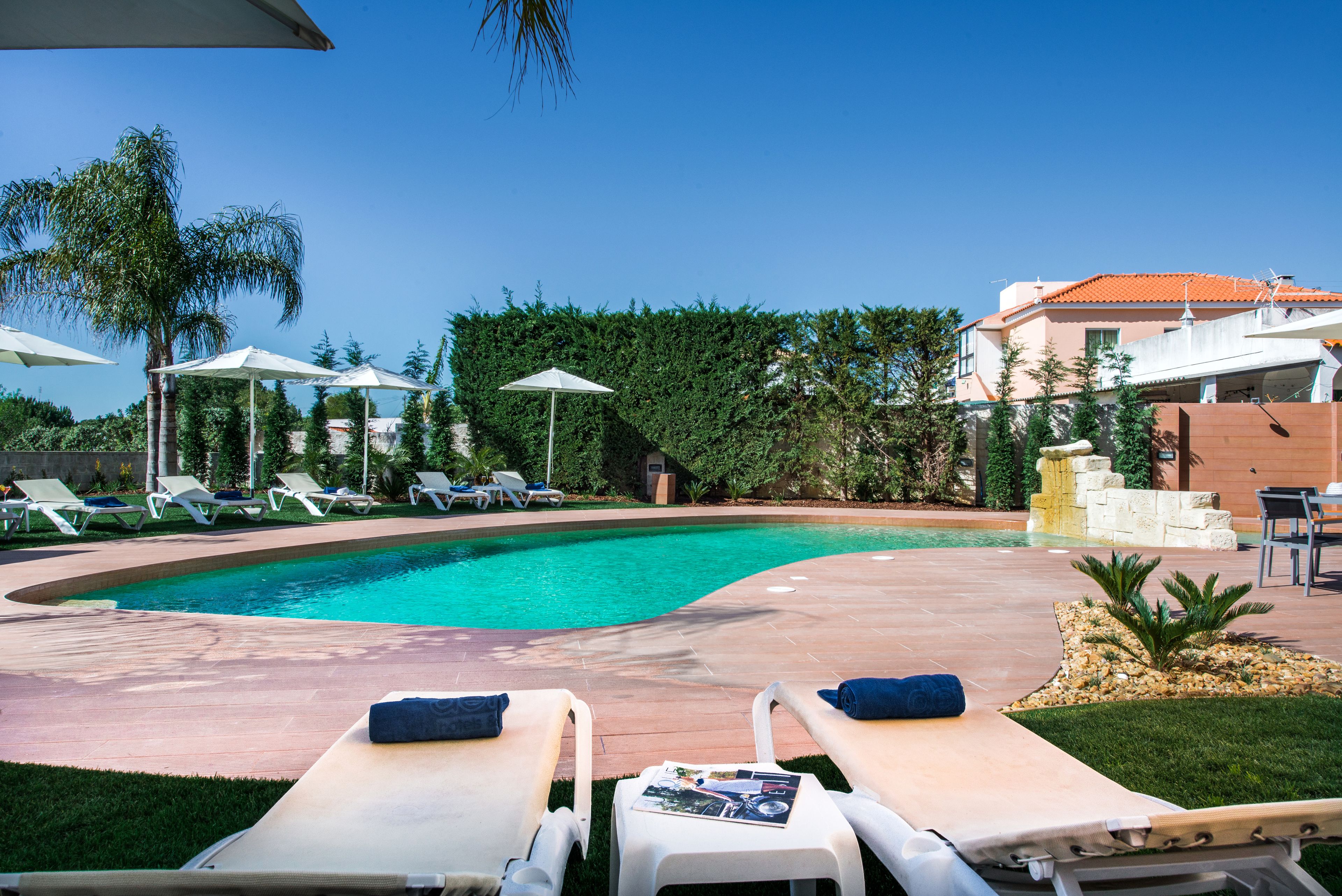 A pool area with lounge chairs and umbrellas, surrounded by greenery and a building with a red roof.