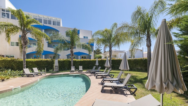 A swimming pool with lounge chairs and umbrellas, surrounded by palm trees and a building with blue accents.