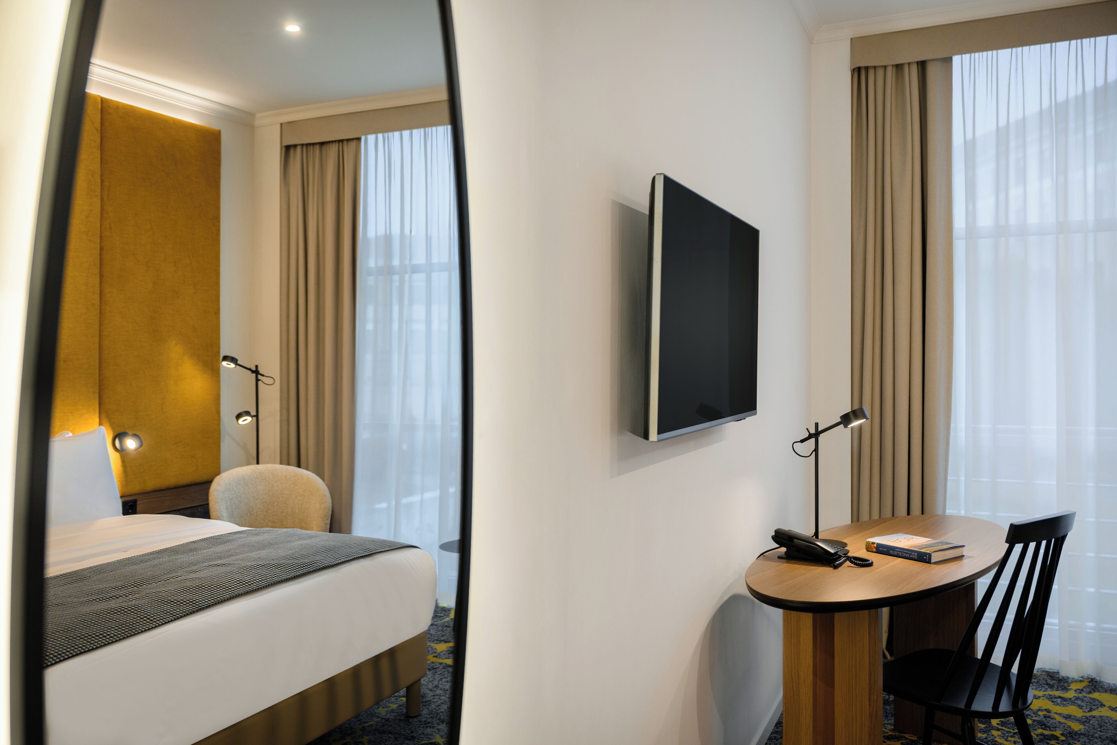 A hotel room with a bed, a desk with a chair, a television, and a window with curtains.
