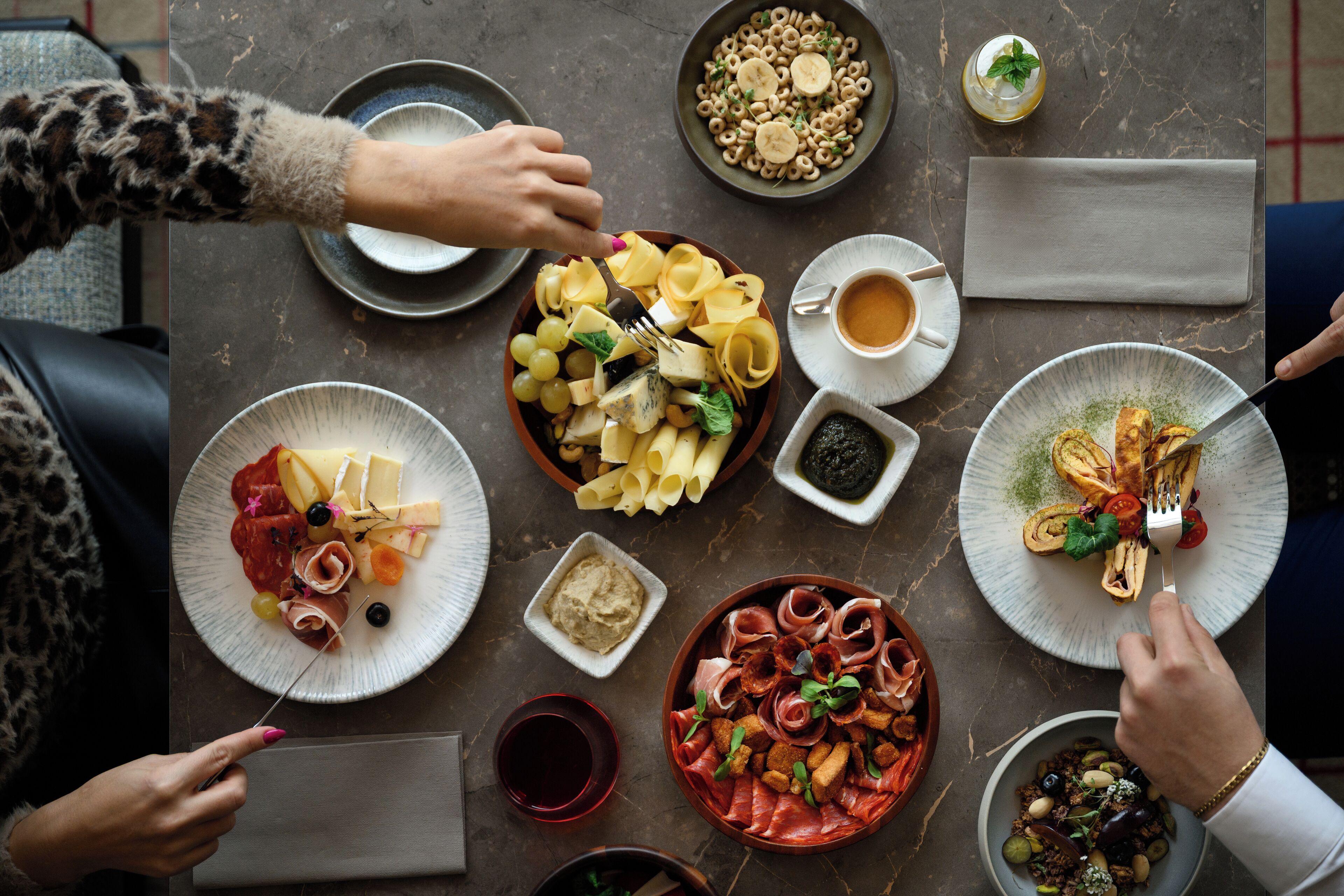 A dining table with various dishes, including pasta, cheese, and meats, surrounded by people.