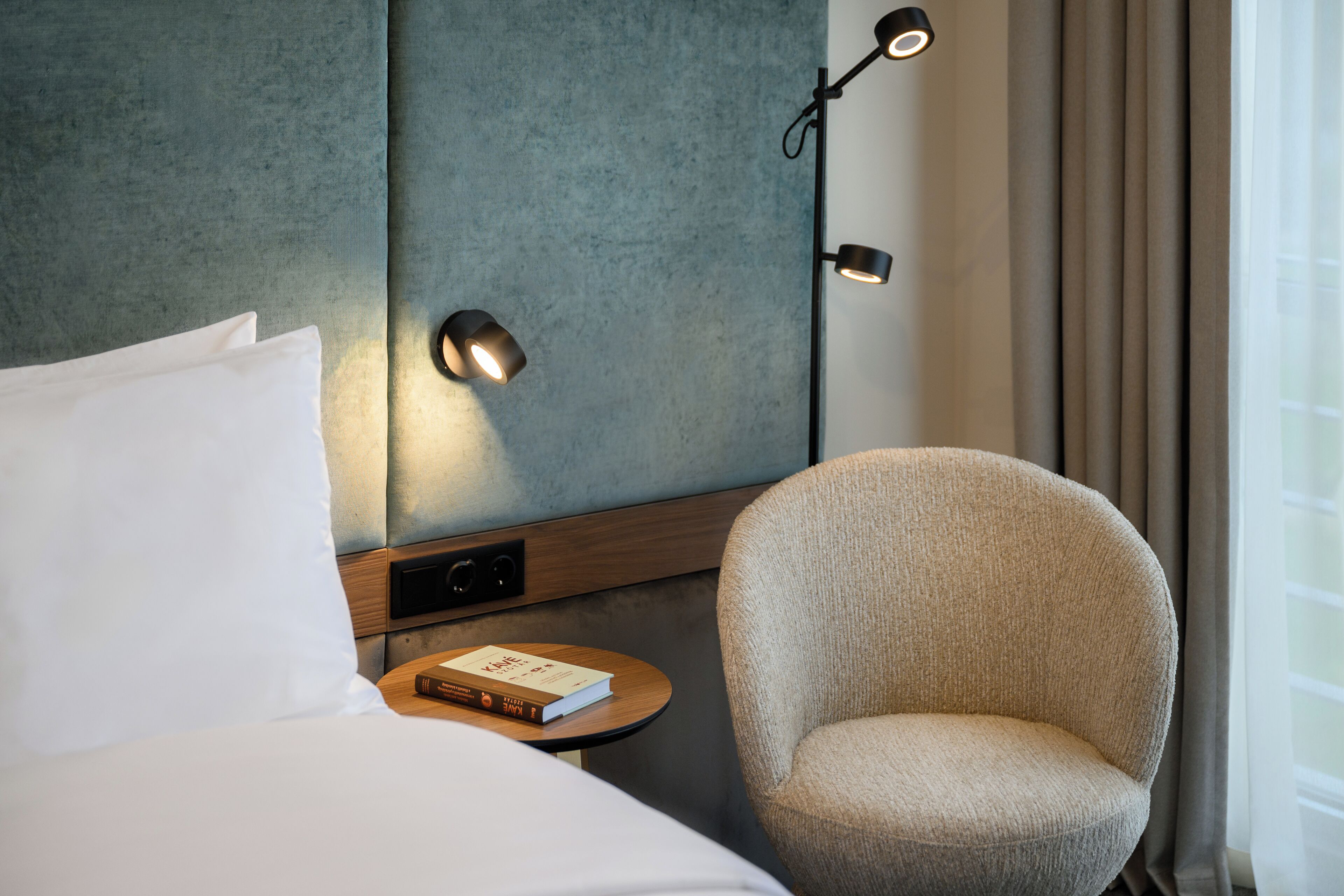 A hotel room with a bed, a chair, a bedside table with a book, and a wall-mounted lamp.