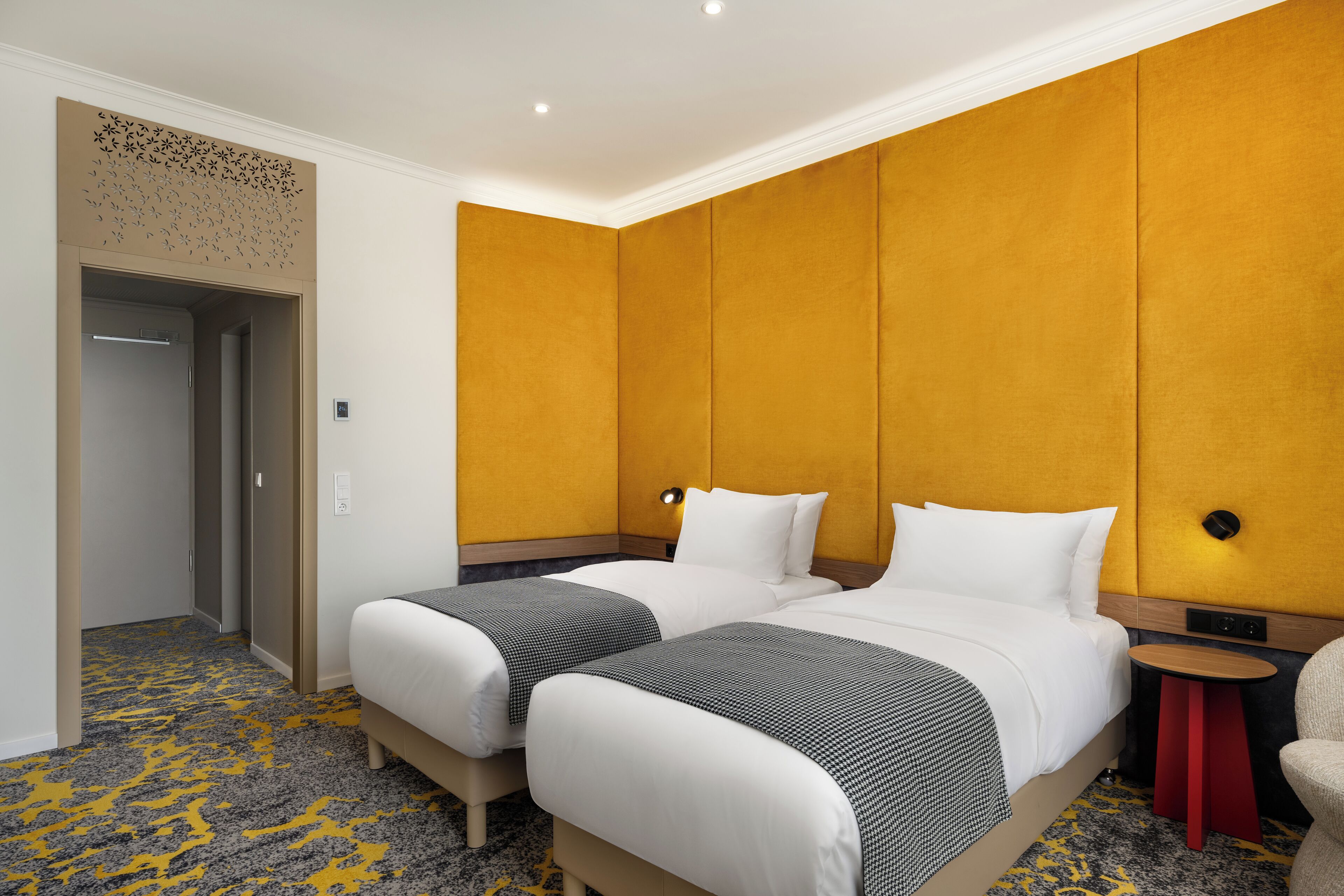 Habitación de hotel con dos camas, una mesita redonda pequeña, una silla y un panel decorativo en la pared.