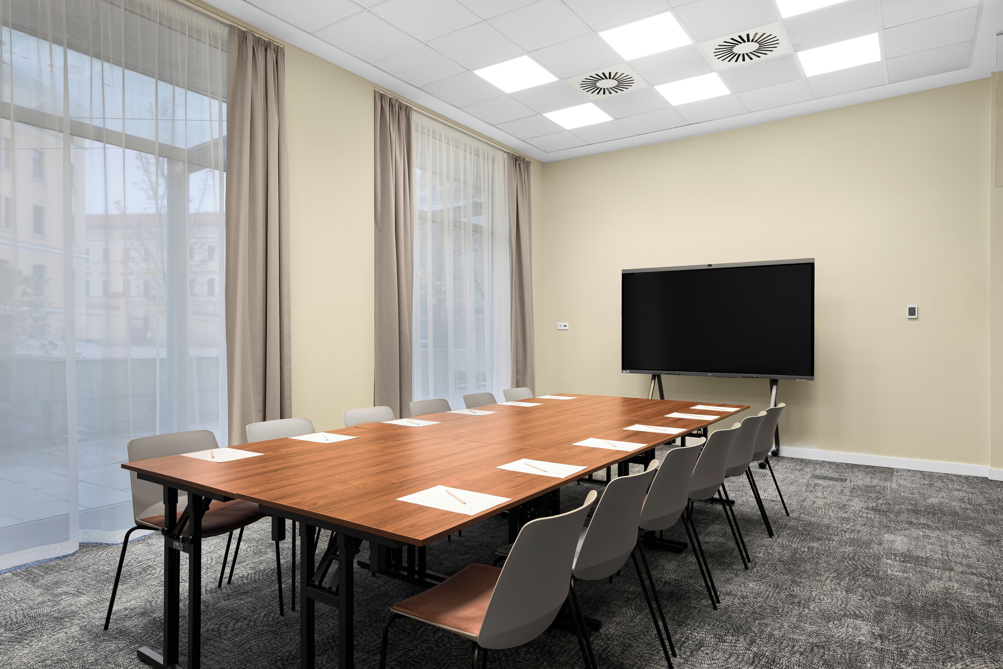 A conference room with a long wooden table, grey chairs, a large screen, and large windows with curtains.