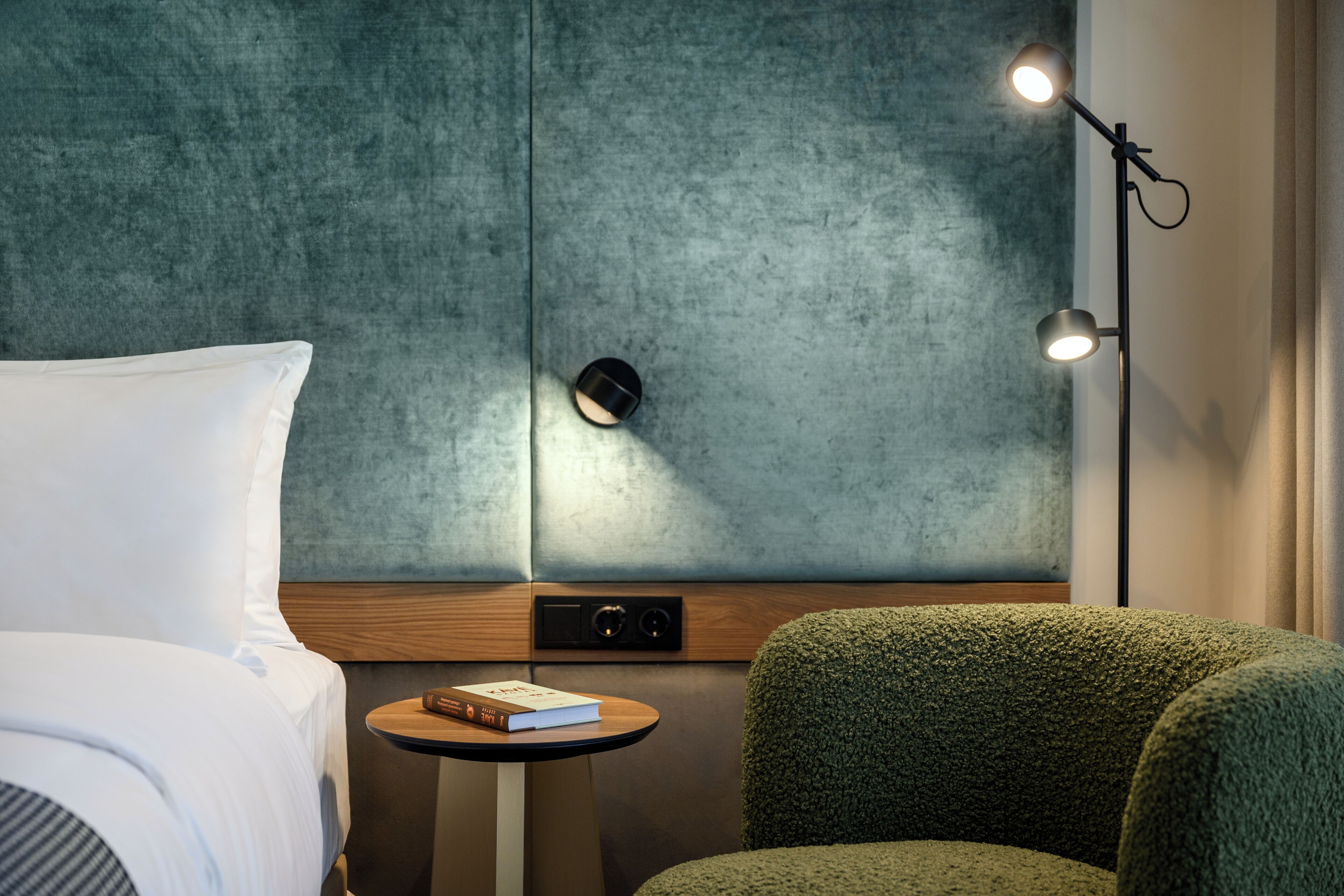 A modern hotel room with a large bed, a green upholstered armchair, a small round table with a book, and a wall-mounted light fixture.