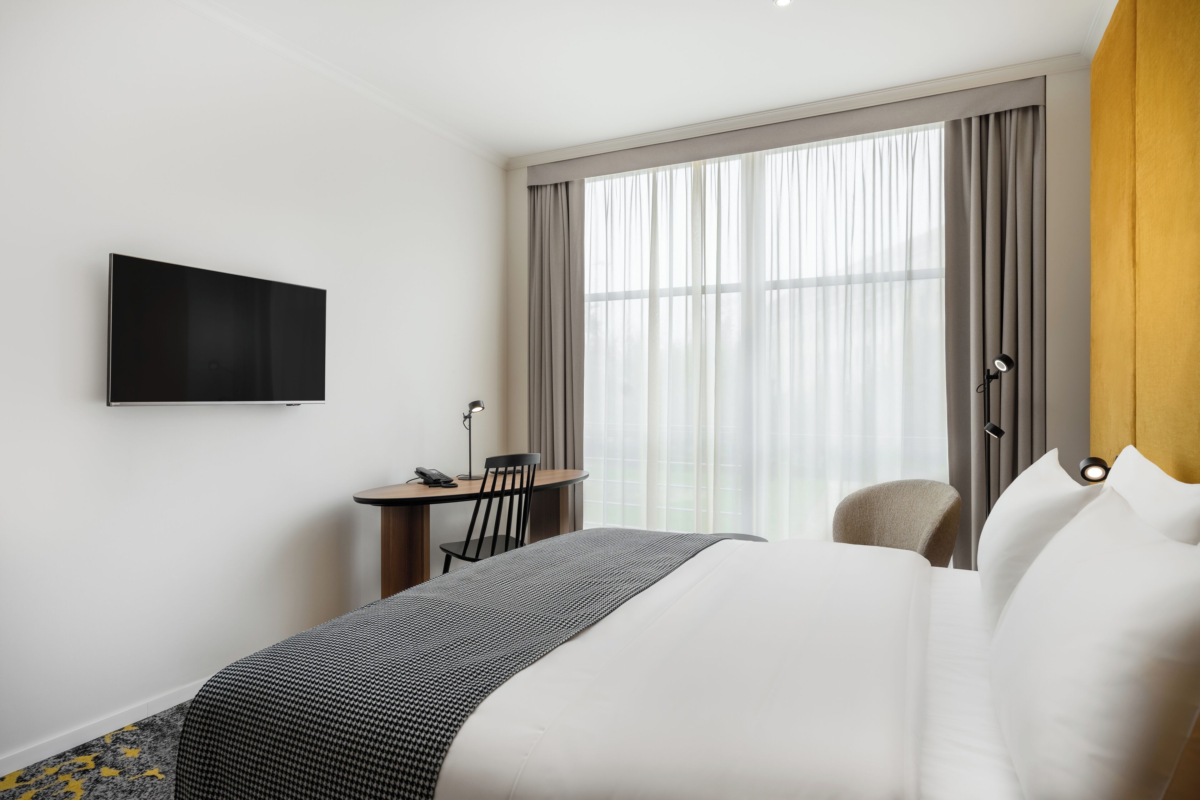 A hotel room with a bed, a desk with a chair, a television on the wall, and a large window with curtains.