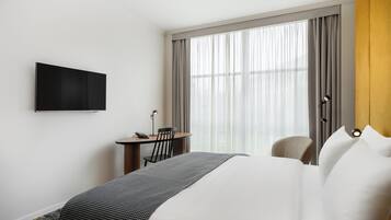 A hotel room with a bed, a desk with a chair, a television on the wall, and a large window with curtains.