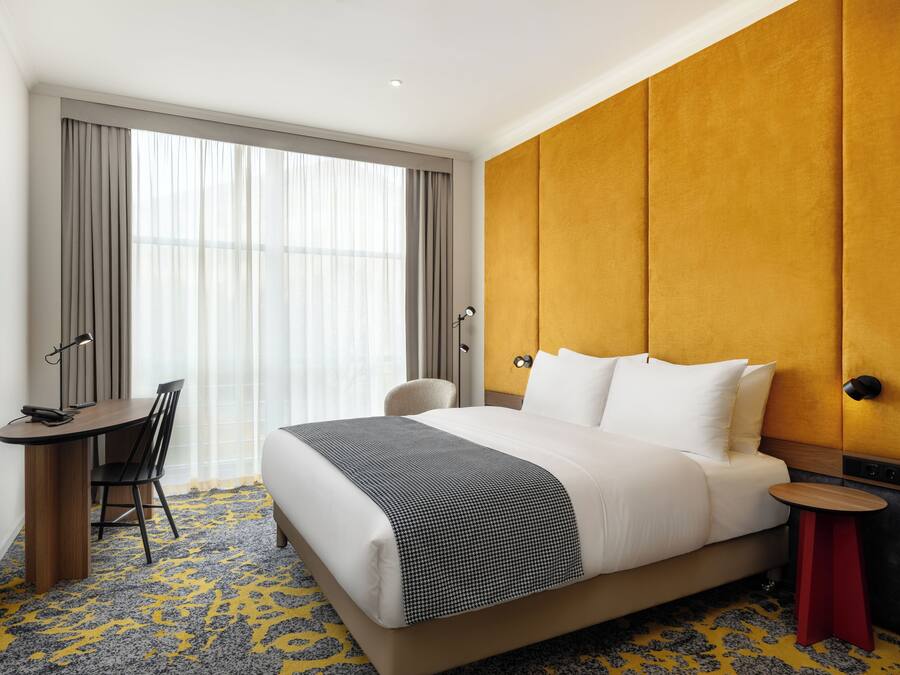 A hotel room with a bed, a desk, a chair, a TV, and a large window with curtains.