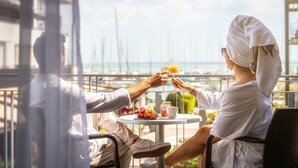 Un couple trinque avec des boissons à une table, avec une vue sur la mer et des bâtiments en arrière-plan.
