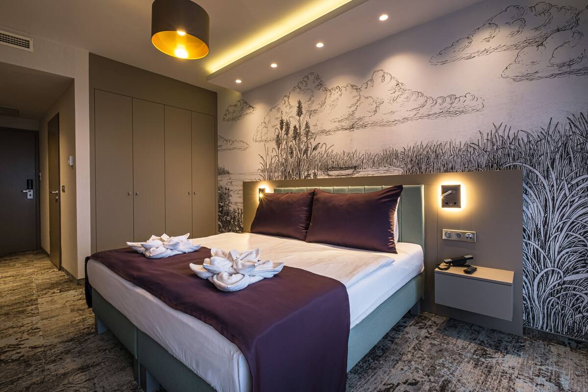 A hotel room with a large bed, a nightstand, a wall mural, and a carpeted floor.