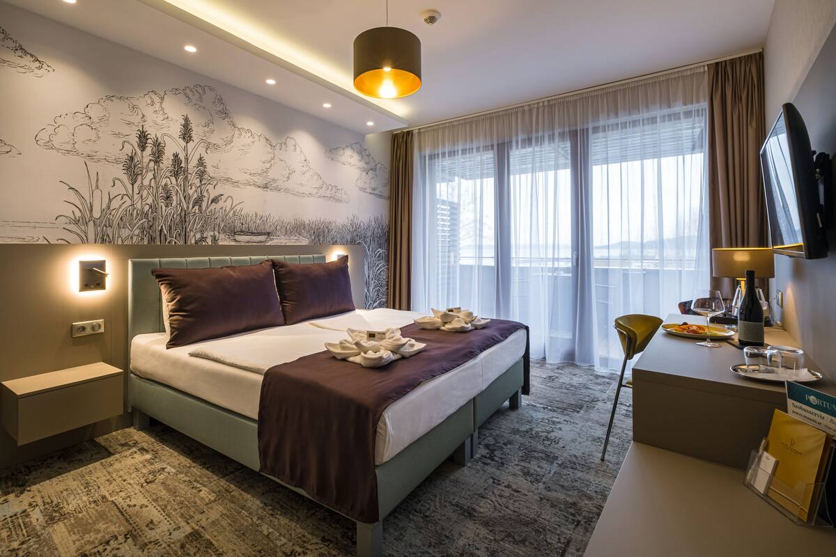 A modern hotel room with a large bed, a desk with a chair, a flat-screen TV, and a large window with curtains.