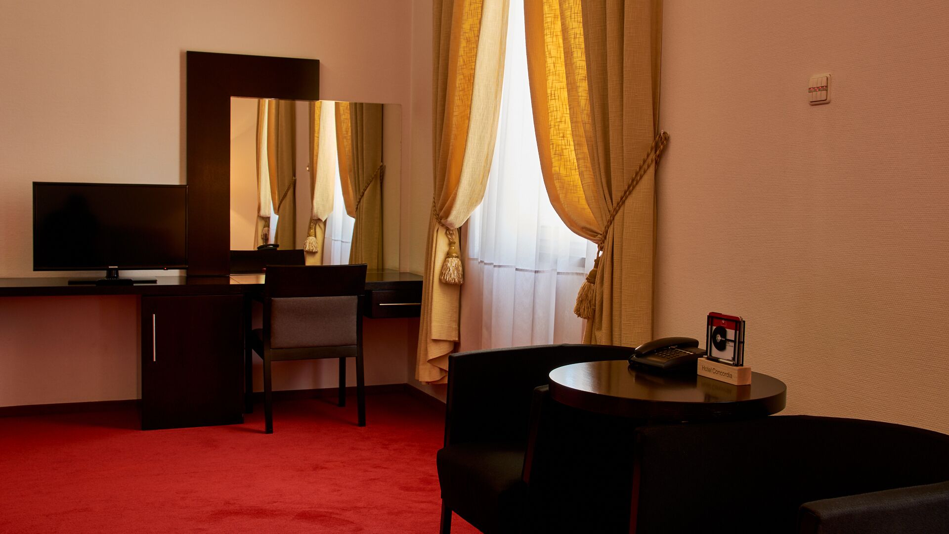 Habitación de hotel con escritorio, silla, televisor y una pequeña mesa con teléfono y portal tarjetas.