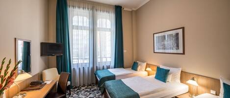 A hotel room with two beds, a desk with a chair, a TV, and a large window with curtains.