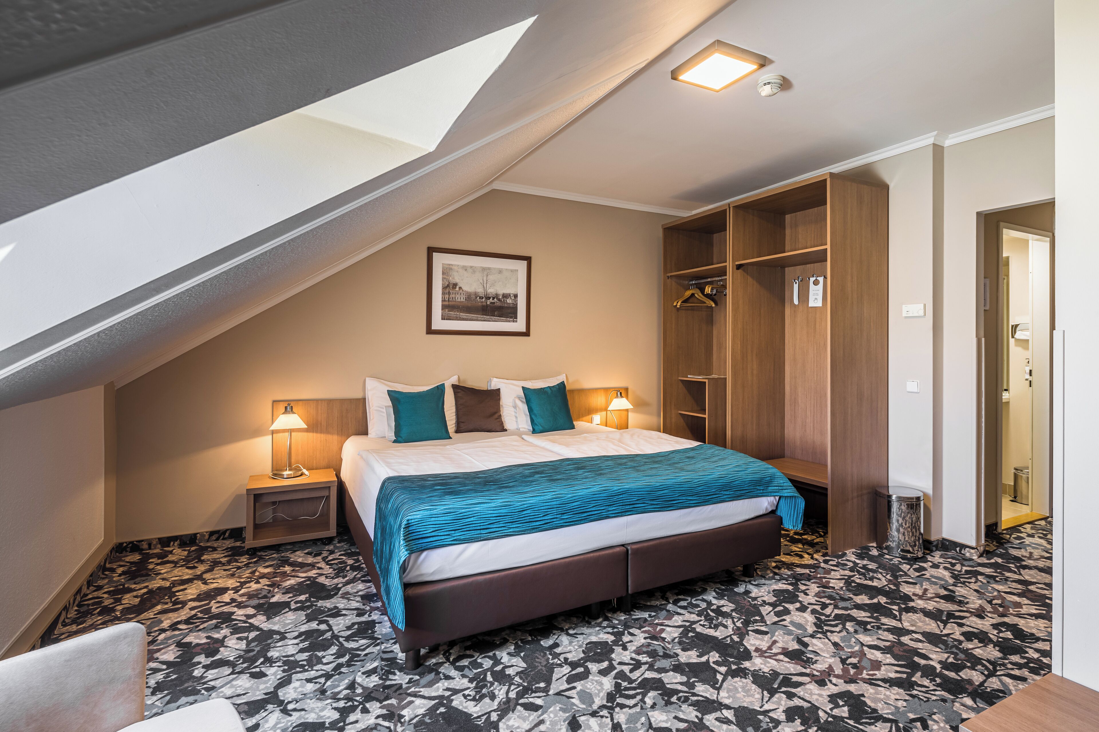 Et hotelværelse med skråvæg, seng med blåt sengetæppe, et natbord med lampe, et træskab og et indrammet billede på væggen.